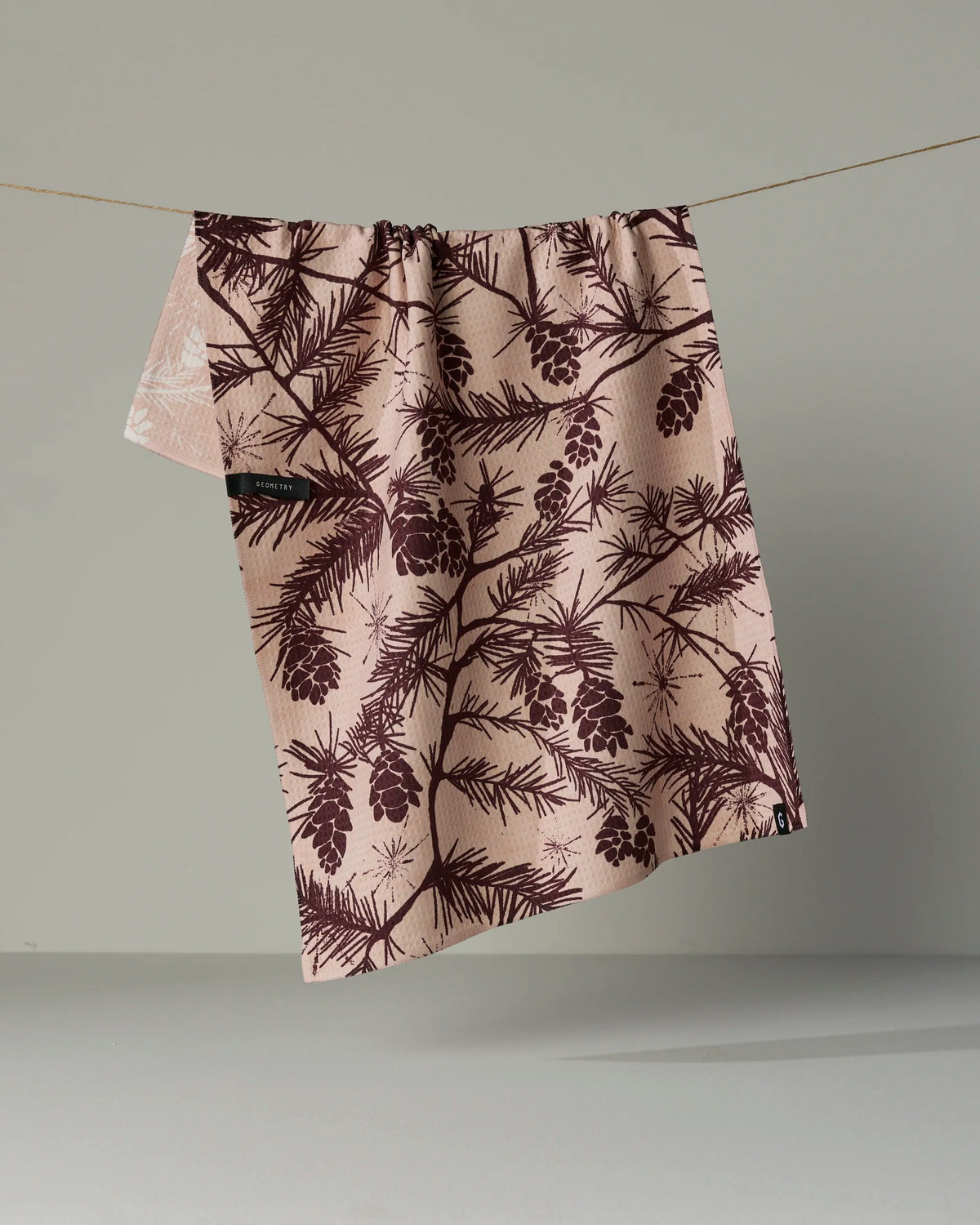 A tea towel from Geometry, called Woodland Calm, hangs on a thin line against a plain, neutral background. Double-sided and adorned with dark brown pinecone and branch motifs on beige fabric, this versatile and stylish piece is both functional and decorative for any kitchen.