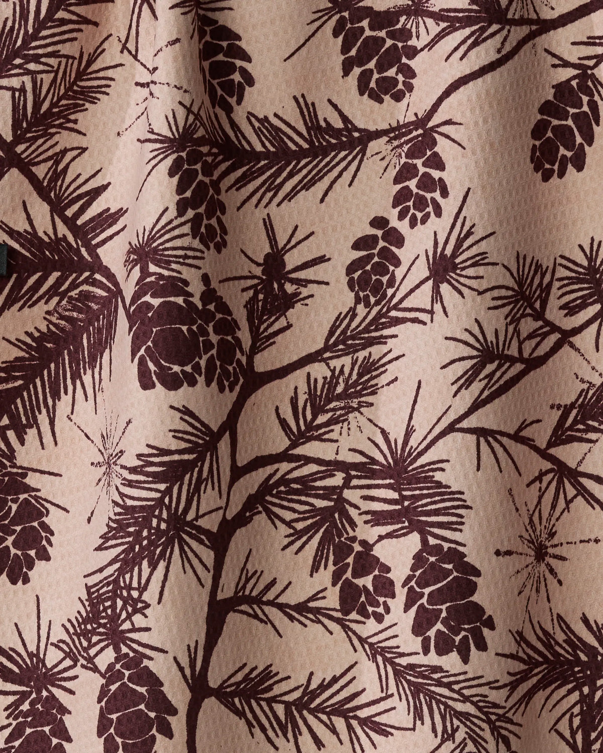 Woodland Calm by Geometry features brown pine tree branches and pinecones printed on light beige textured recycled fabric, with soft shadows suggesting gentle folds—making this kitchen towel both charming and eco-friendly.