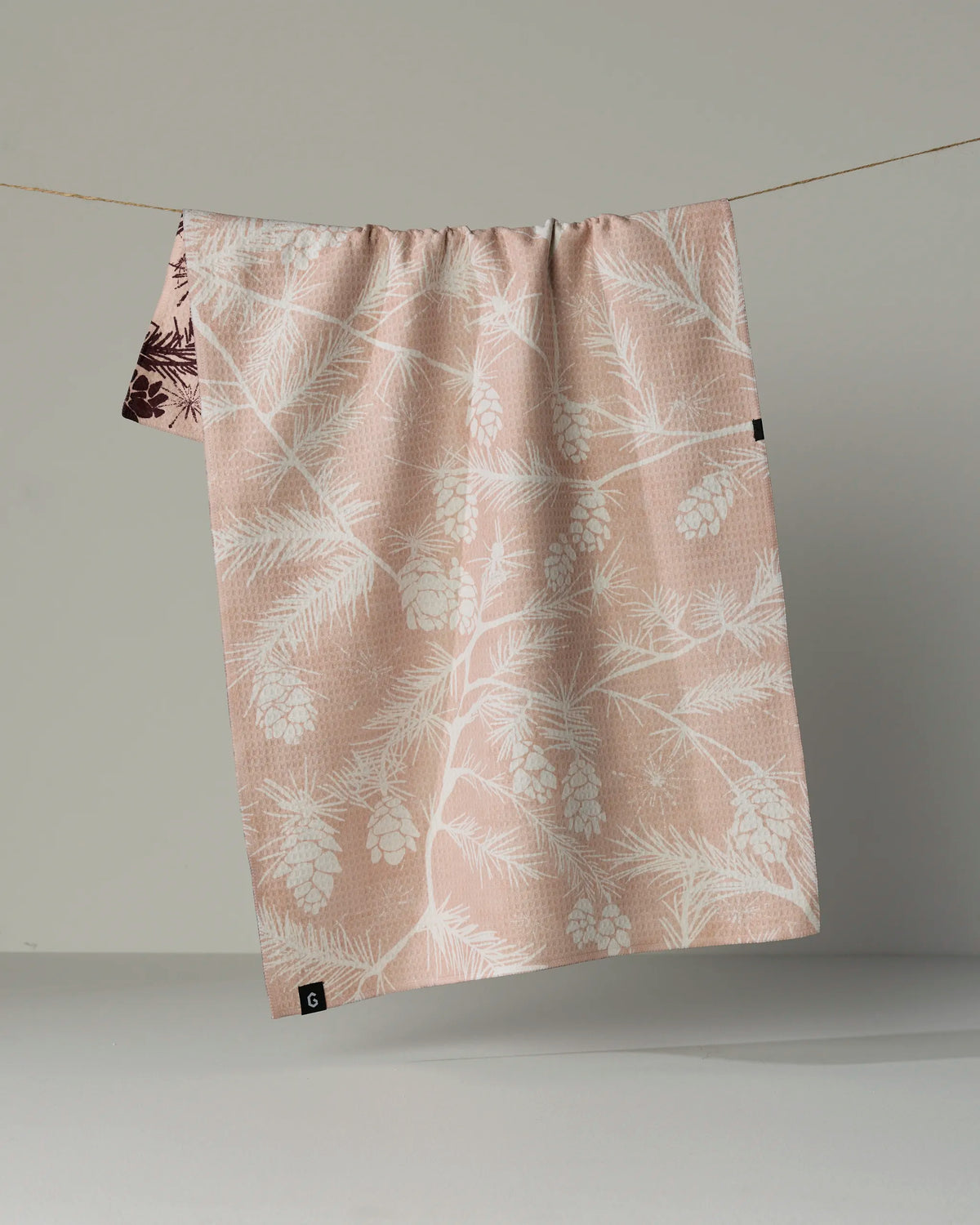 A pale pink, quick-drying tea towel from Geometry called “Woodland Calm,” featuring a white pinecone and pine branch pattern, hangs on a clothesline against a neutral background.