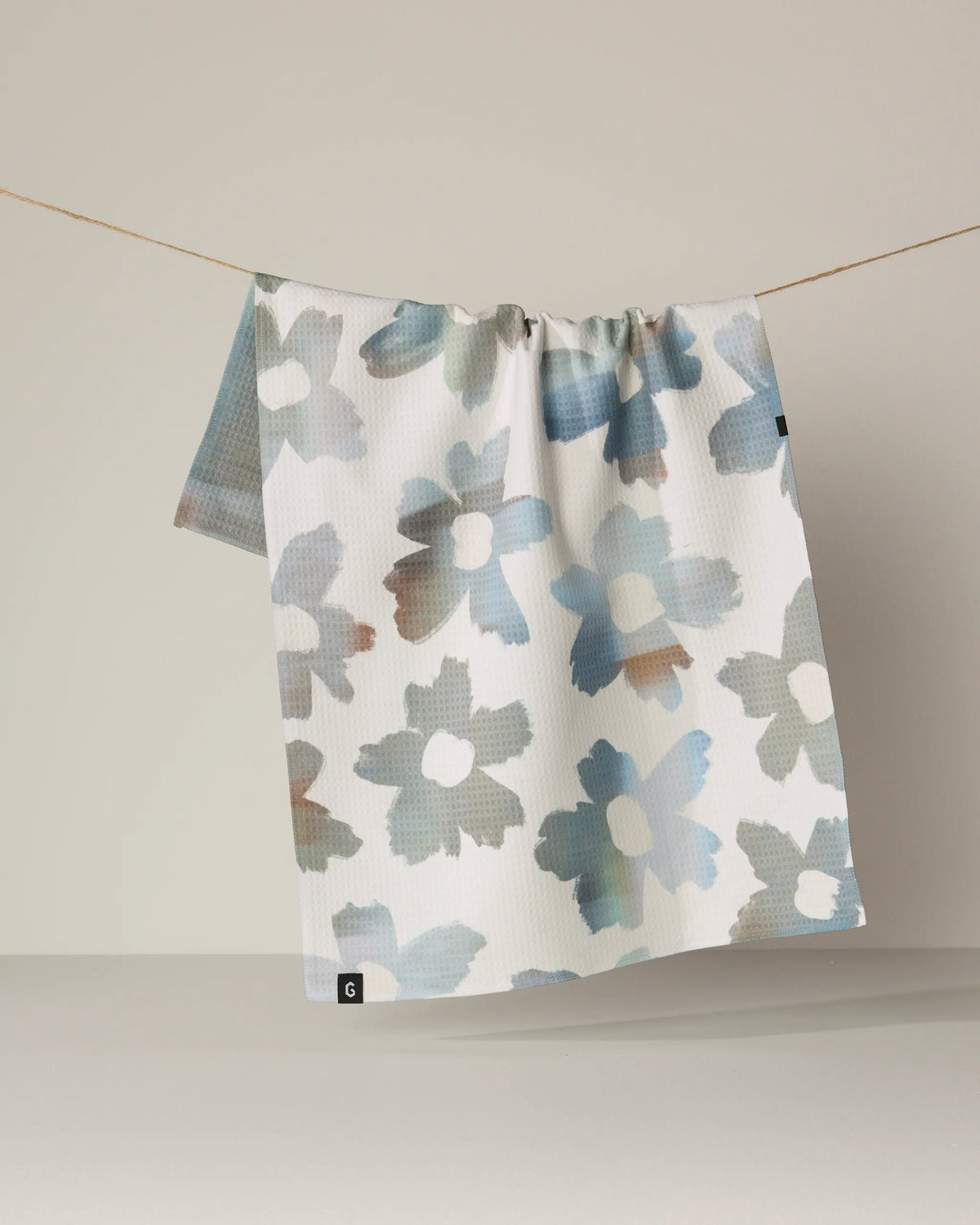 A “Brushed” kitchen towel by Geometry, featuring a blue and brown floral pattern on recycled white fabric, hangs on a thin clothesline against a plain beige background.