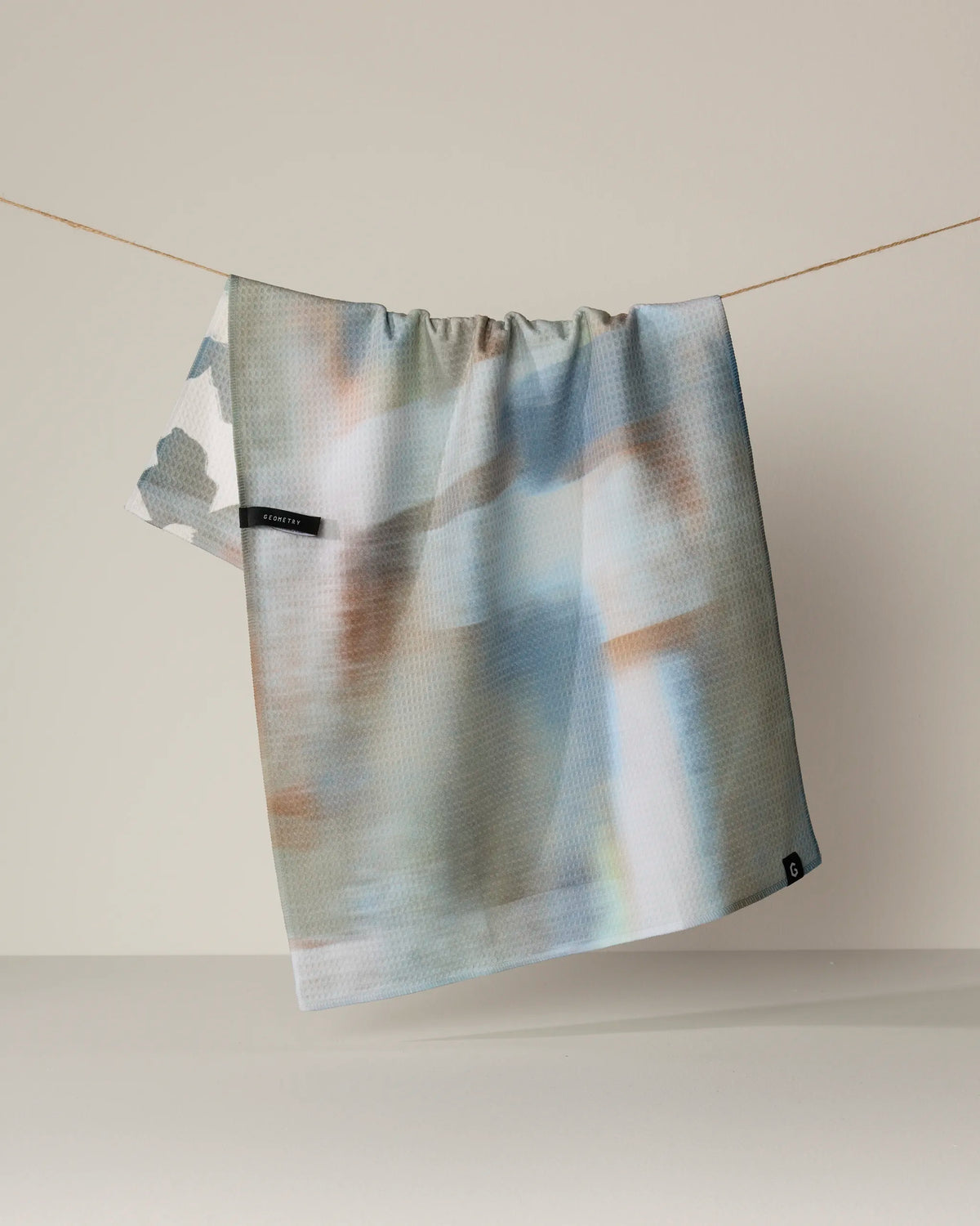 The Geometry Brushed tea towel, featuring double-sided abstract patterns in pastel hues, hangs on a string against a plain, light background.