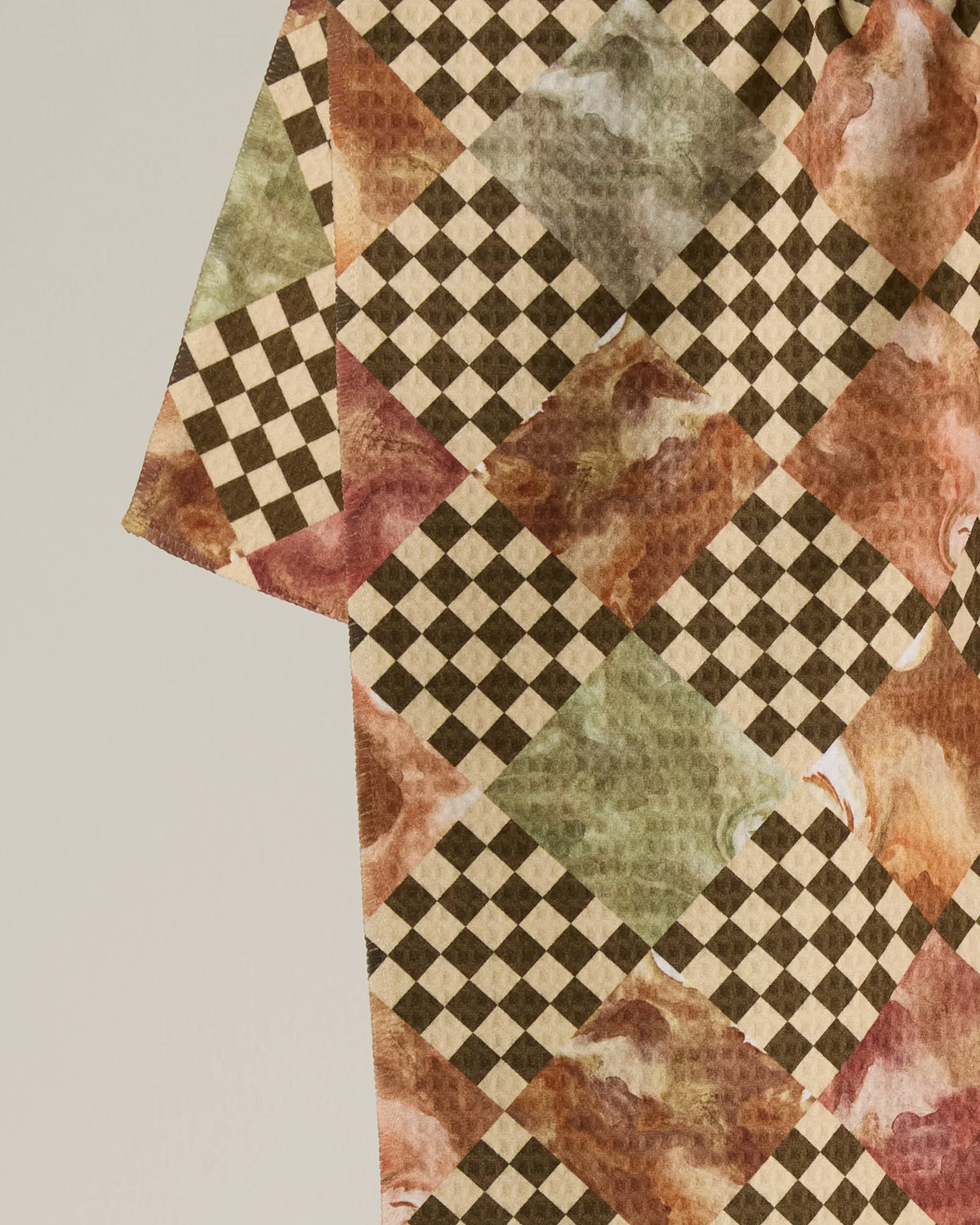 A close-up of the “Check” tea towel by Geometry, showcasing patterned fabric with alternating brown checkered squares and marbled squares in green, red, and beige on a light background—an ideal choice for a recycled kitchen towel or absorbent tea towel that blends sustainability with style.