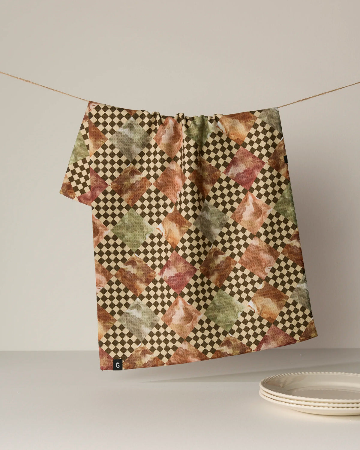 A double-sided “Check” tea towel by Geometry, featuring diamond and checkered designs in earthy tones, hangs on a string above a stack of white plates.