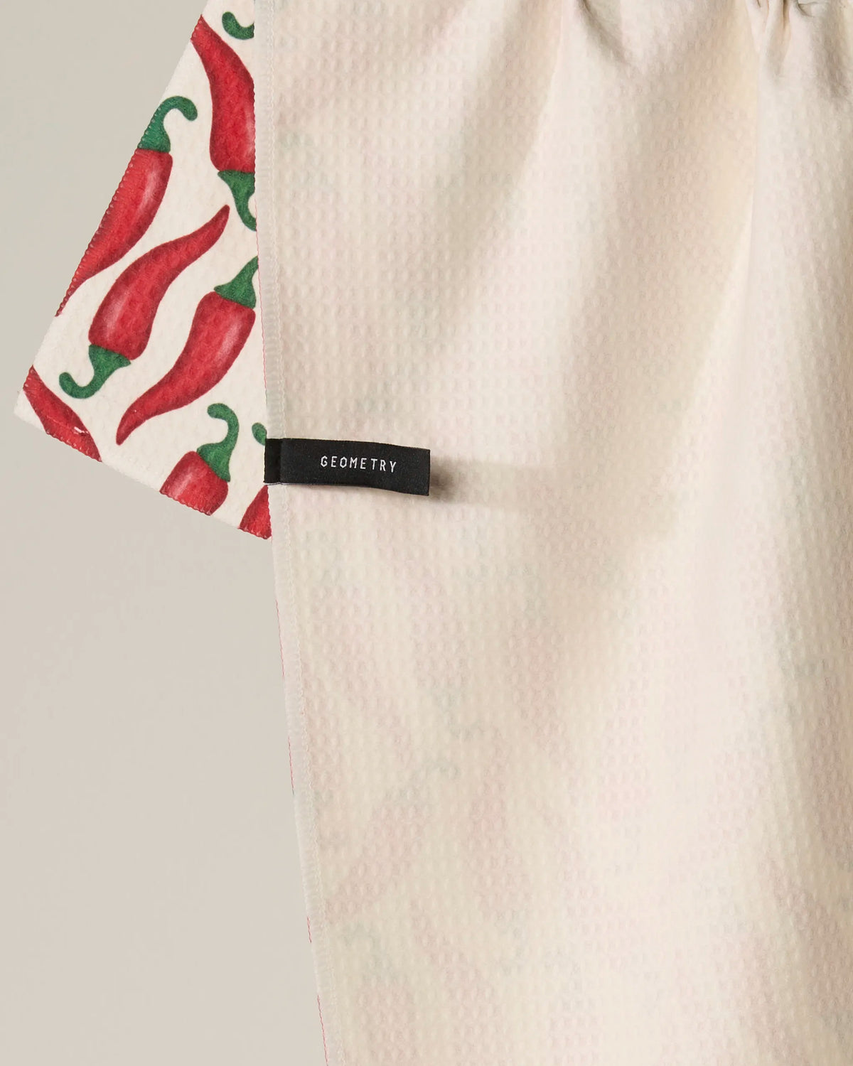 The Geometry “Chili” towel features a striking red chili pepper print along one edge and a black tag bearing the Geometry brand, while the majority of the absorbent, quick-drying towel remains a simple light shade—perfect for adding personality and everyday practicality to your kitchen.