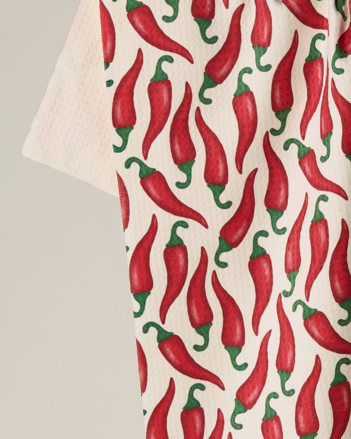A close-up of the Geometry “Chili” double-sided tea towel, showcasing a repeating pattern of red chili peppers with green stems on a white background.