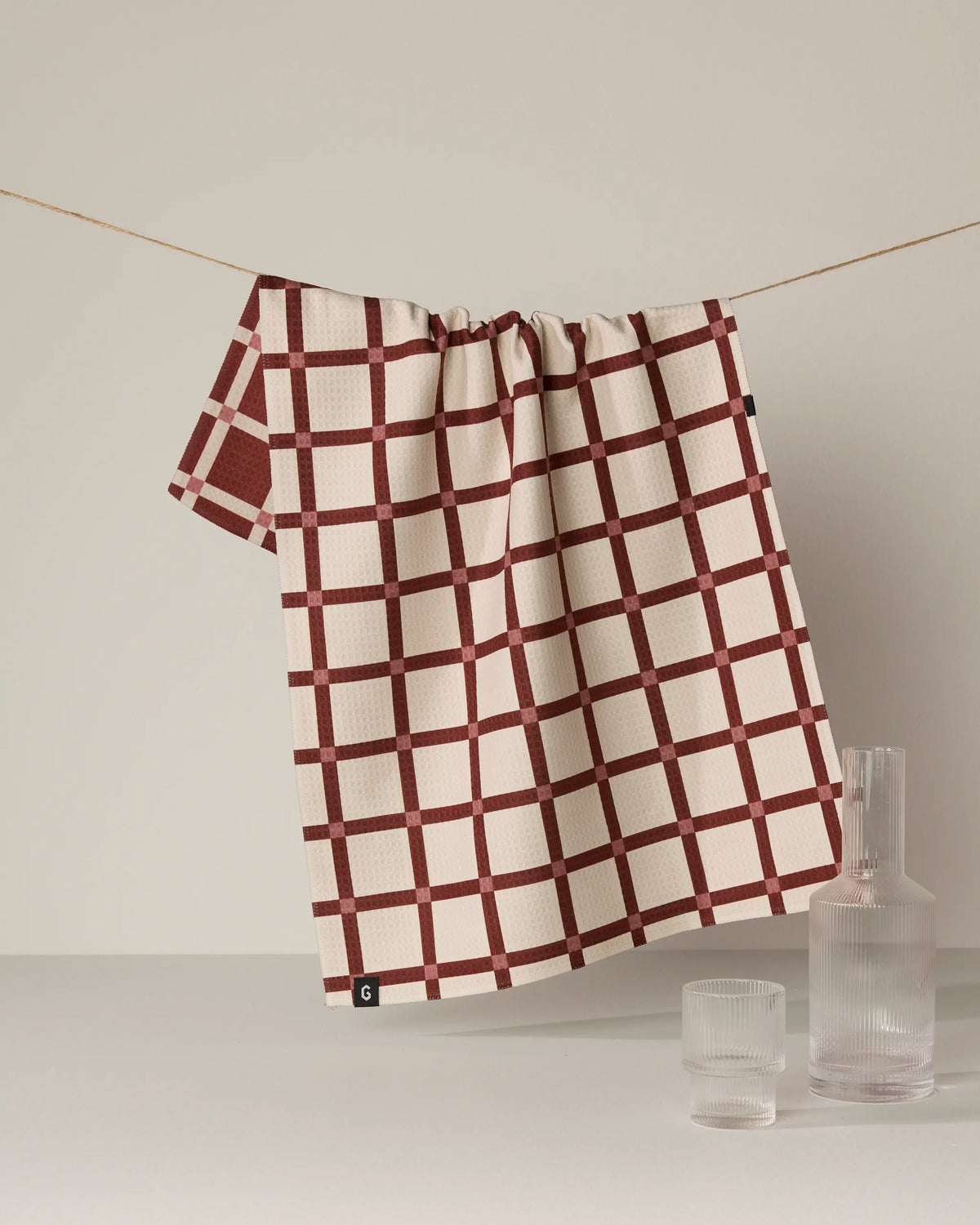 A quick-drying Geometry Clay Tile tea towel, featuring a cream and red checkered pattern, hangs on a string above a white surface, with a clear glass carafe and two matching glasses nearby.