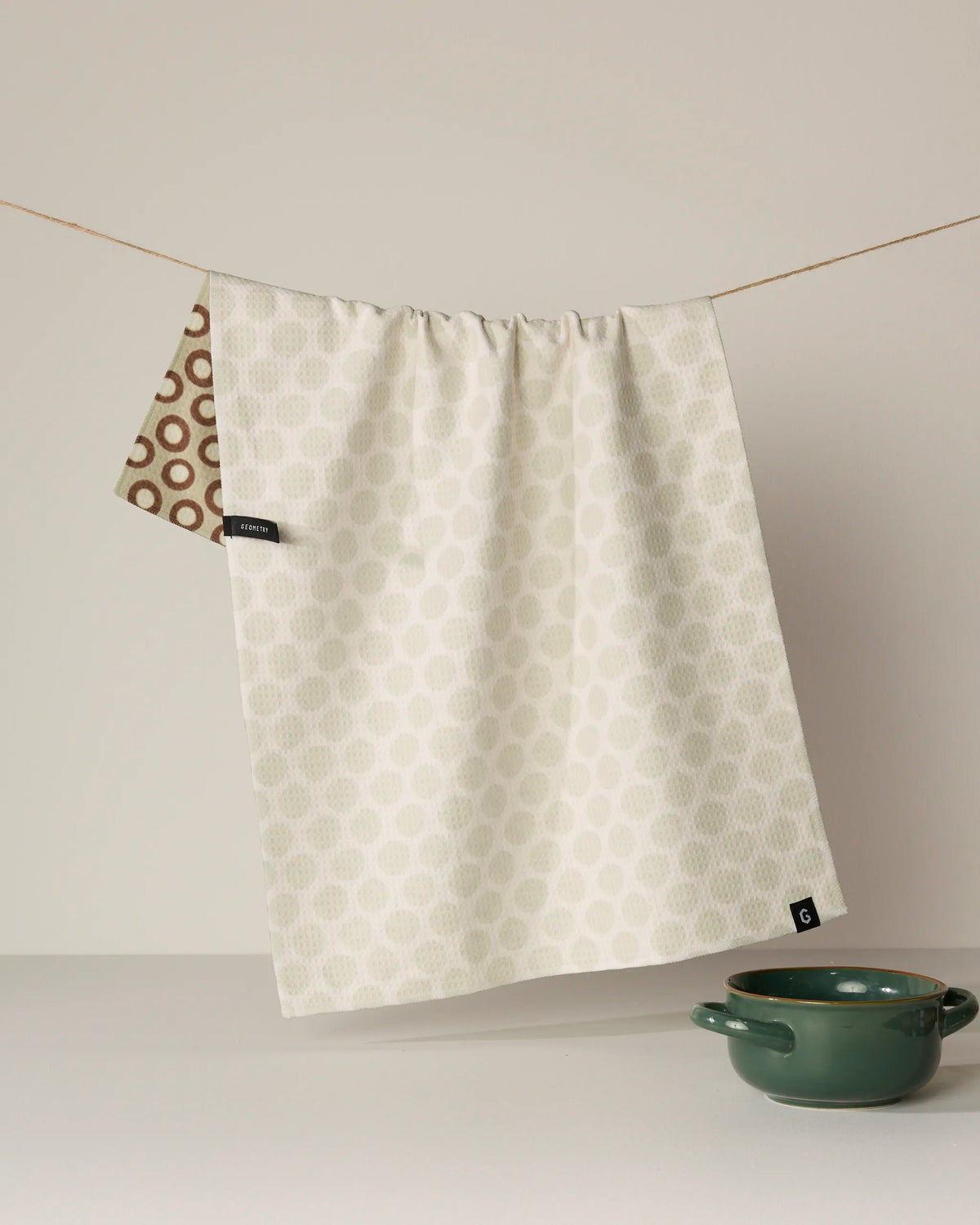 A cream double-sided tea towel from Geometry, named Dotty, with a subtle dot pattern hangs on a line. A section with brown pattern peeks out, and a green bowl rests nearby on a white surface. Crafted from recycled materials, the Dotty tea towel is absorbent, quick-drying, stylish, and eco-friendly.