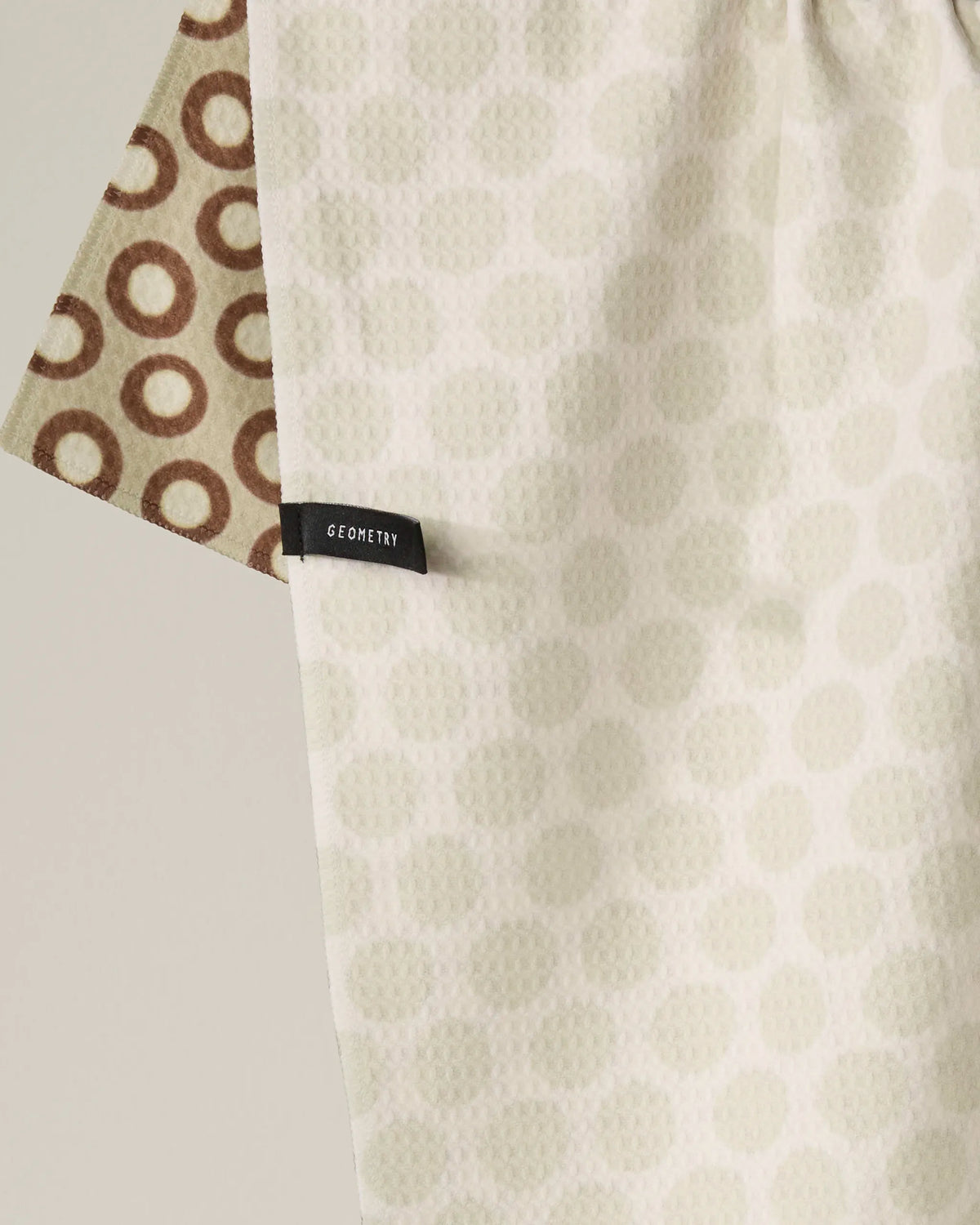 Close-up of two textured fabric pieces—one cream-colored with light dots and the other brown with circular patterns. A black tag reading Geometry is attached to the cream fabric. “Dotty” by Geometry is a double-sided tea towel made from recycled materials, thoughtfully blending aesthetic appeal with eco-friendly functionality.