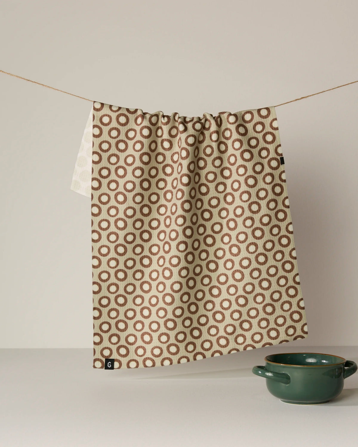 A beige double-sided “Dotty” tea towel by Geometry, featuring a pattern of brown circles, hangs from a string. Below, a green ceramic bowl rests on a light surface against a neutral background. Made from recycled materials, this absorbent and quick-drying towel offers both sustainability and style for your space.