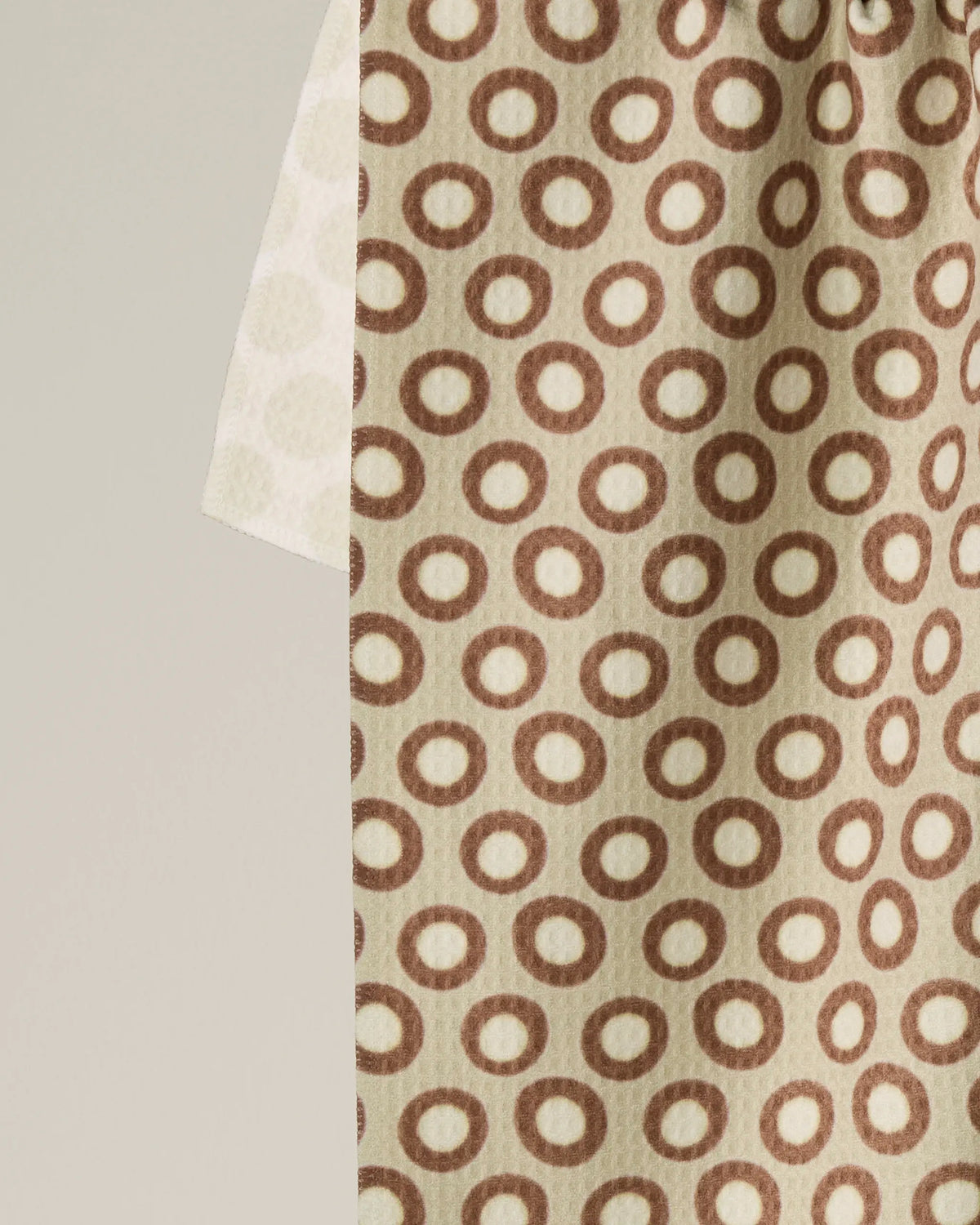 A beige double-sided tea towel, Dotty by Geometry, featuring a repeating pattern of brown circles and made from recycled materials, hangs partially overlapping another similar lighter-toned fabric against a plain background. Both towels are absorbent, quick-drying, and eco-friendly.