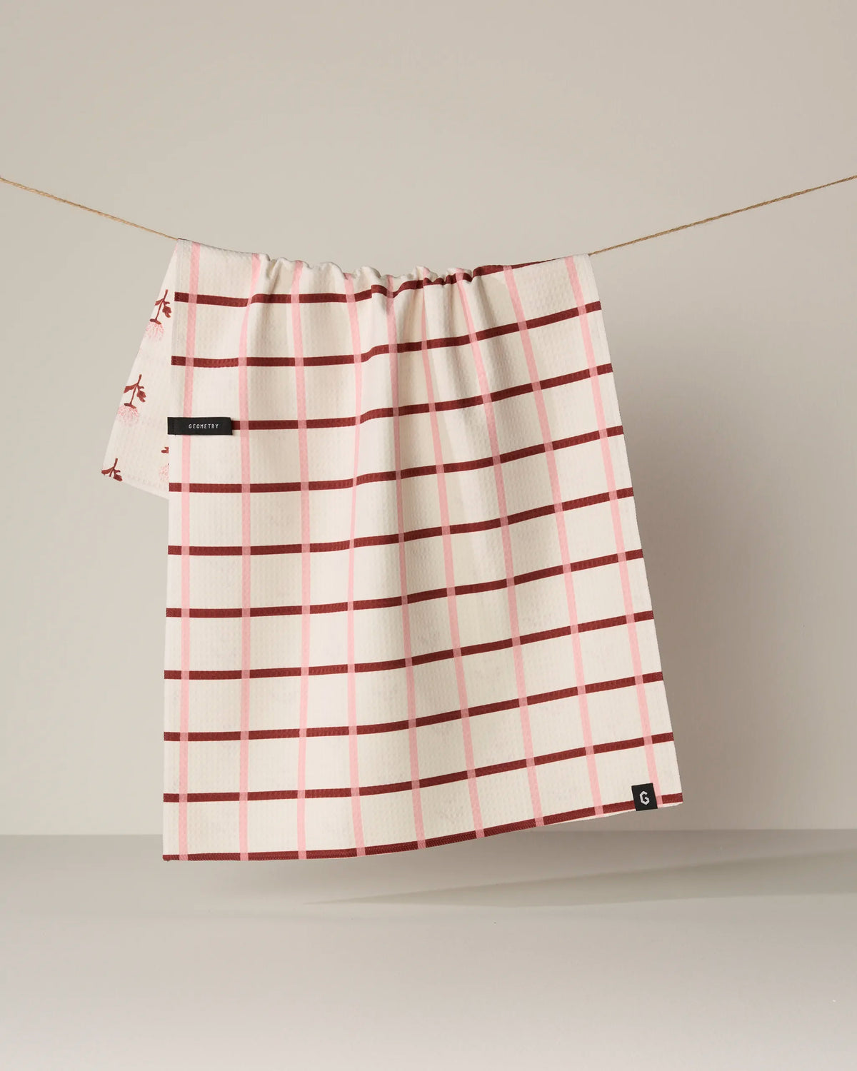 The Geometry Fleur tea towel in white with a red checkered pattern is displayed double-sided, hanging on a string against a plain beige background.