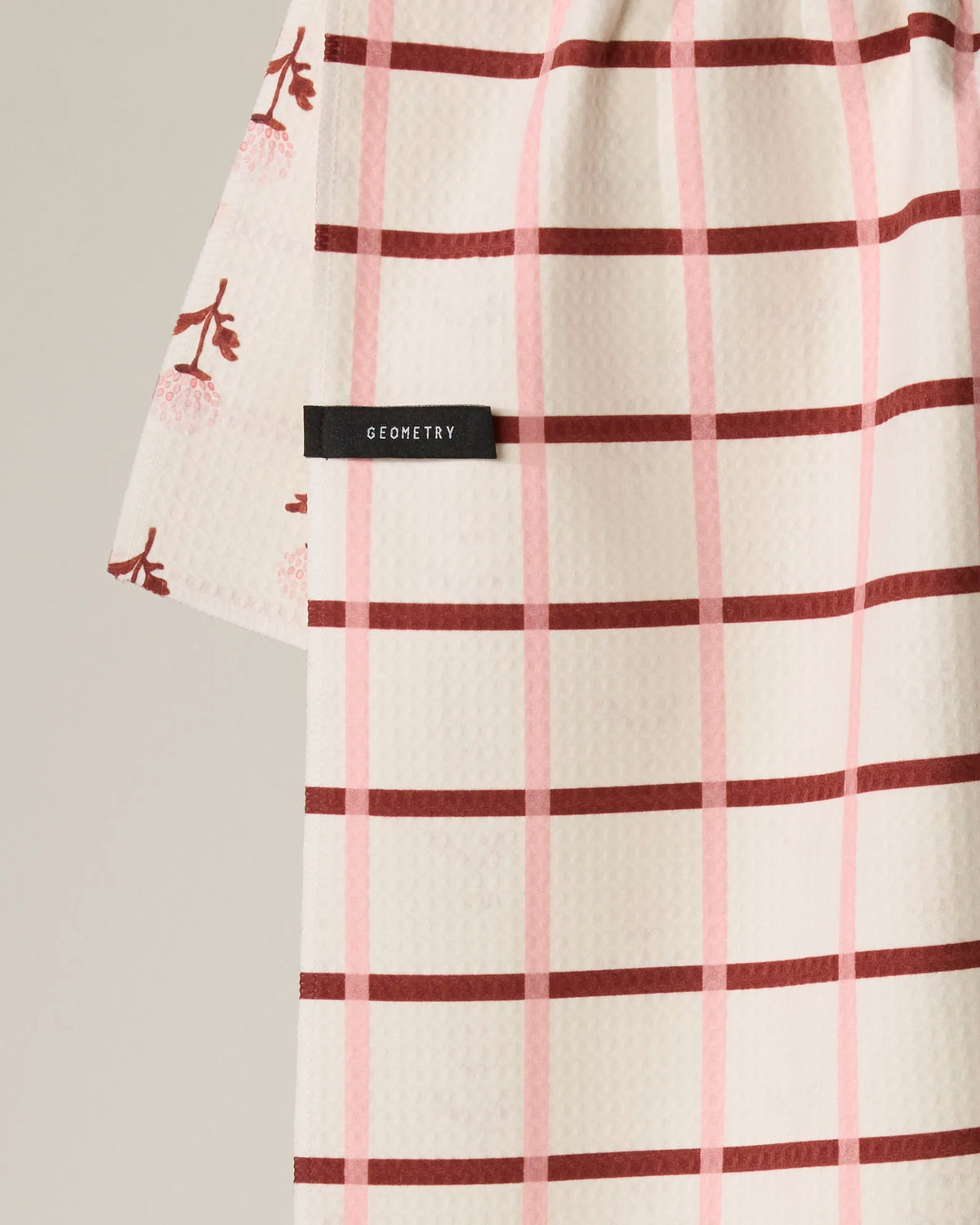 The Fleur tea towel by Geometry is a white, textured towel featuring a pink and burgundy plaid pattern, with a small black GEOMETRY tag and a corner adorned with a pink floral design. Made from quick-drying recycled materials, it’s both practical and stylish.