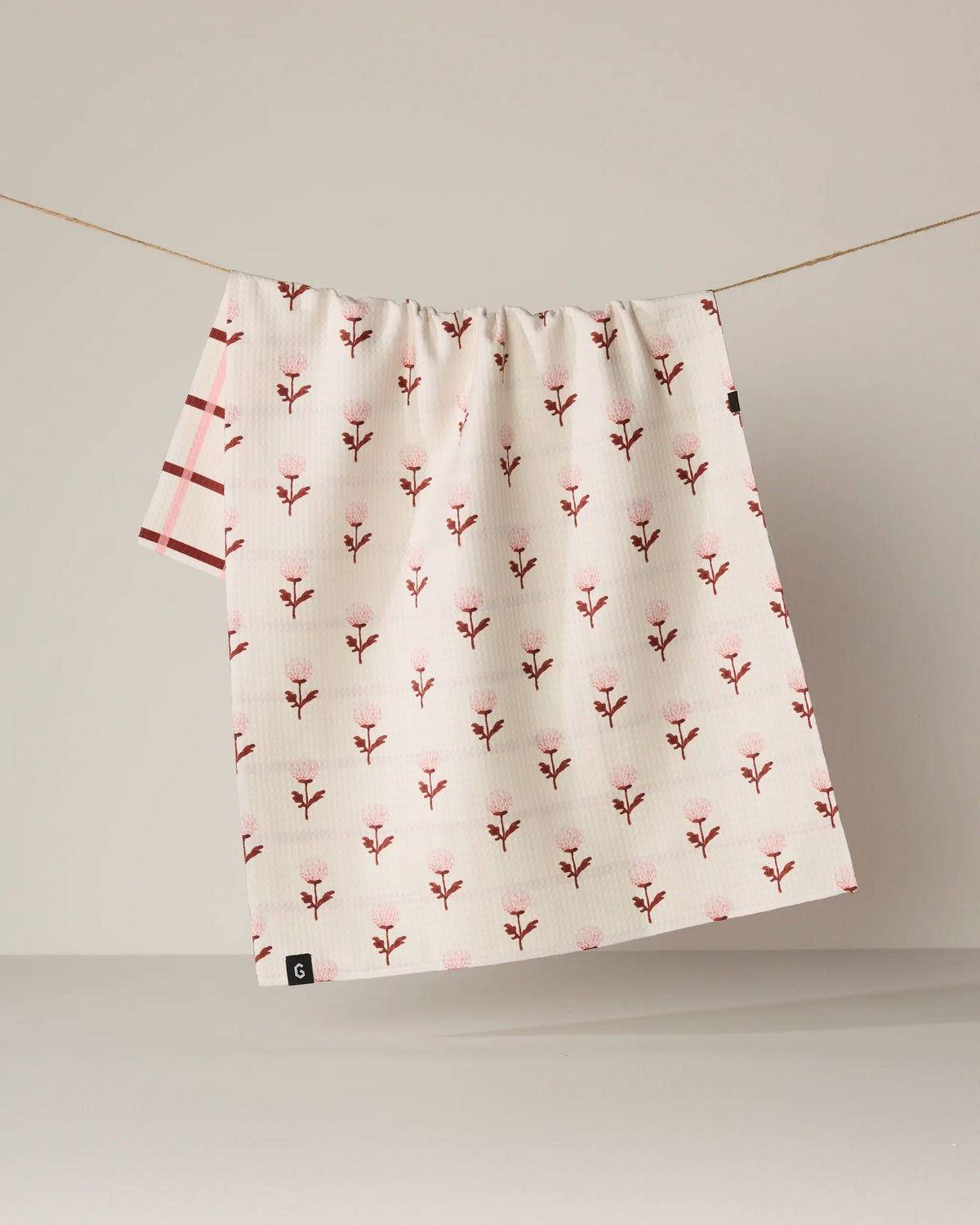 Two Geometry “Fleur” tea towels hang on a line. One double-sided towel features delicate pink floral motifs, while the other showcases a pink checkered design. Both quick-drying Geometry Fleur towels are displayed against a plain beige background.