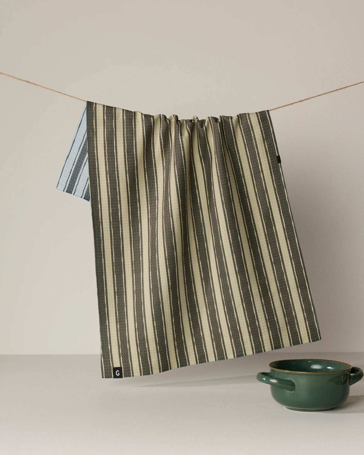 A double-sided Flow tea towel by Geometry, featuring striking stripes, hangs on a string against a neutral background, while a green ceramic bowl rests on the surface below.