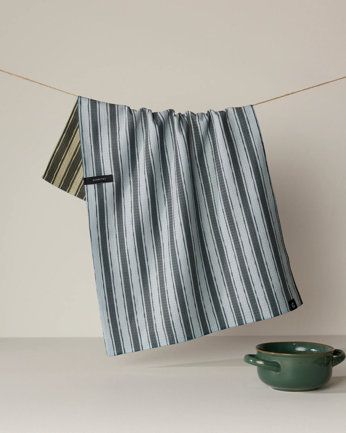 A Geometry Flow quick-drying tea towel, featuring blue and white stripes, hangs on a clothesline above a green ceramic bowl on a light surface with a neutral background.