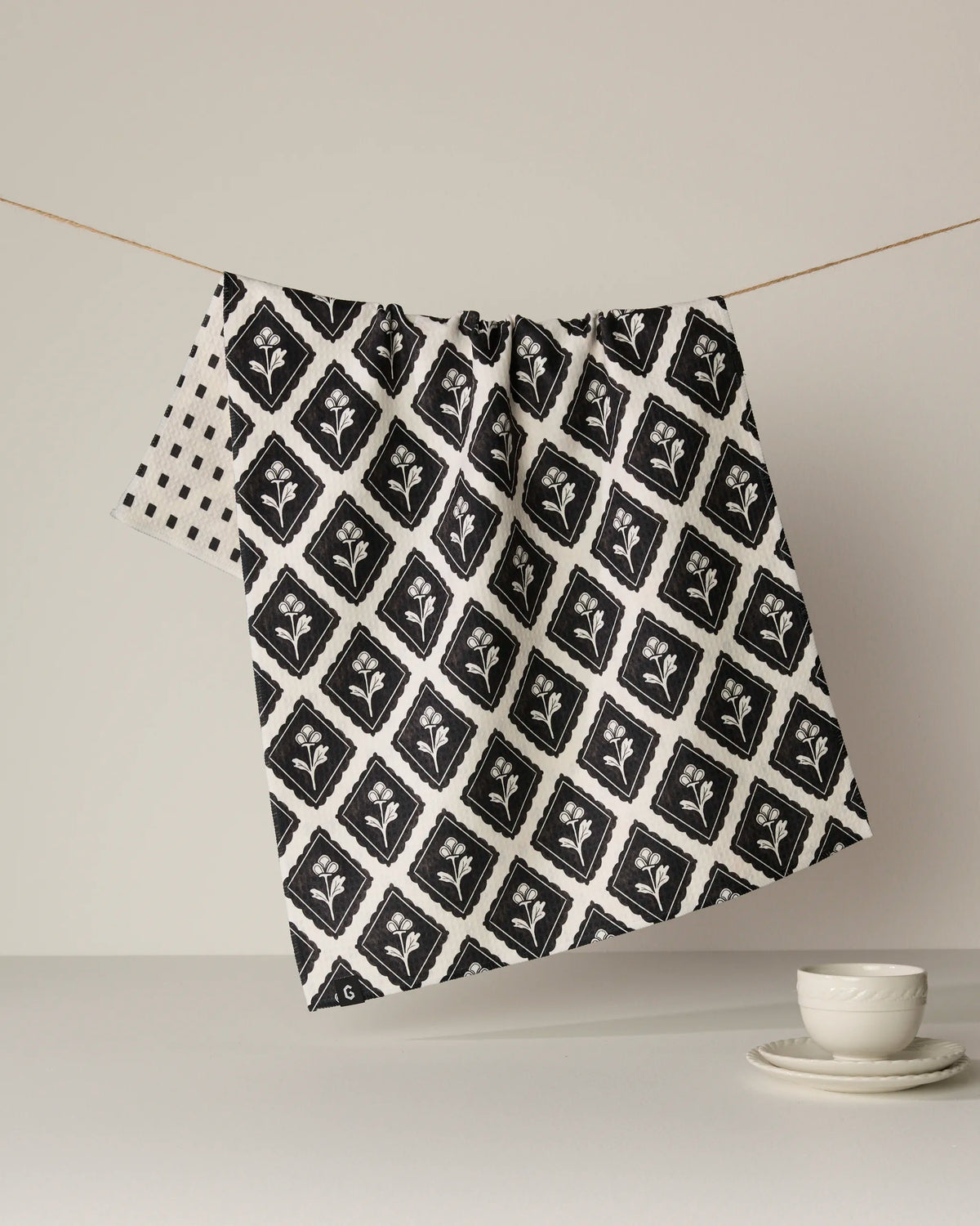 A double-sided Geometry “Maggie” tea towel featuring a black and white pattern hangs on a string against a neutral background, with a white cup and saucer placed on the surface in the foreground.