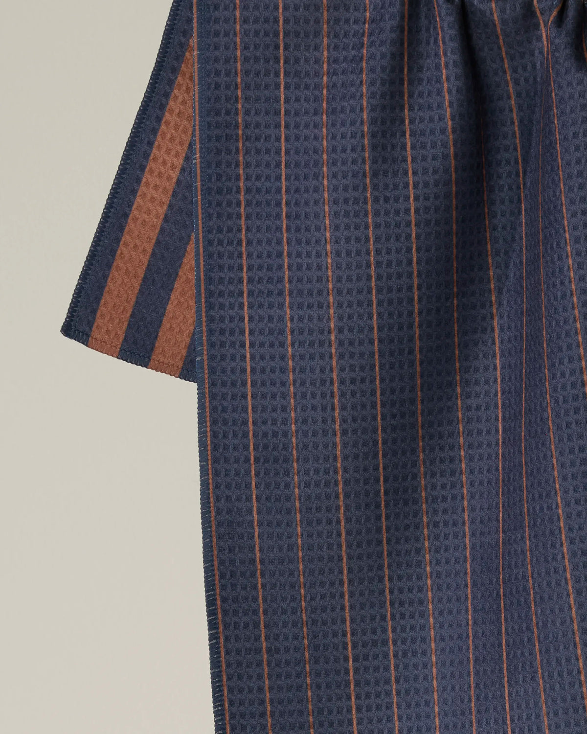 A close-up of Geometry’s Midnight Stripe fabric reveals its dark blue hue with thin brown vertical stripes and textured pattern, showcasing part of a sleeve against a light background—evocative of the refined weave found in a double-sided tea towel.
