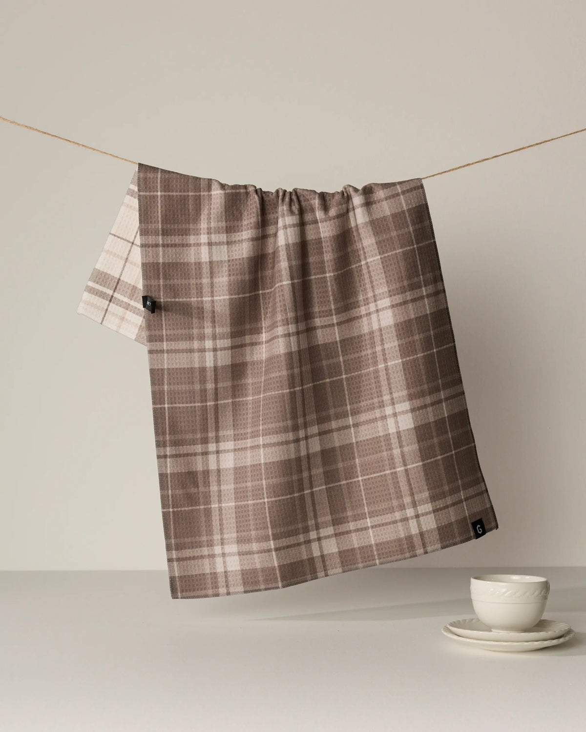 An Oat Plaid absorbent quick-drying towel by Geometry, featuring a brown and beige plaid pattern, hangs on a string against a neutral background, with a stacked white cup and saucer set positioned on the white surface below.