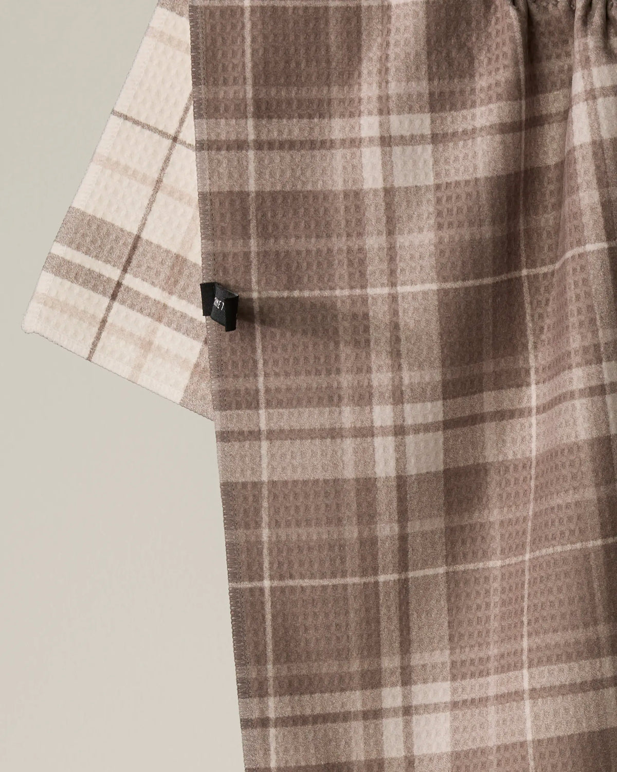 Close-up of two layered Oat Plaid fabrics by Geometry in shades of beige and brown, with a small black fabric tag on one layer. The intersecting stripes form a checkered design—making this double-sided tea towel both stylish and practical.