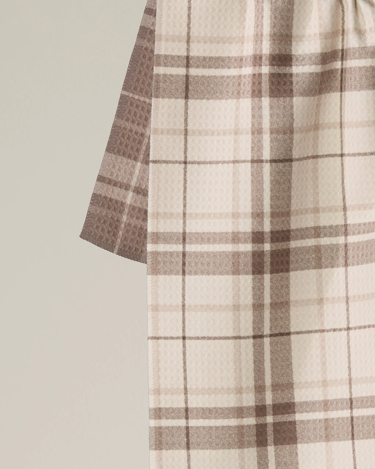 A close-up of the Oat Plaid kitchen towel by Geometry, featuring a beige and brown plaid pattern, draped vertically against a plain, light background.
