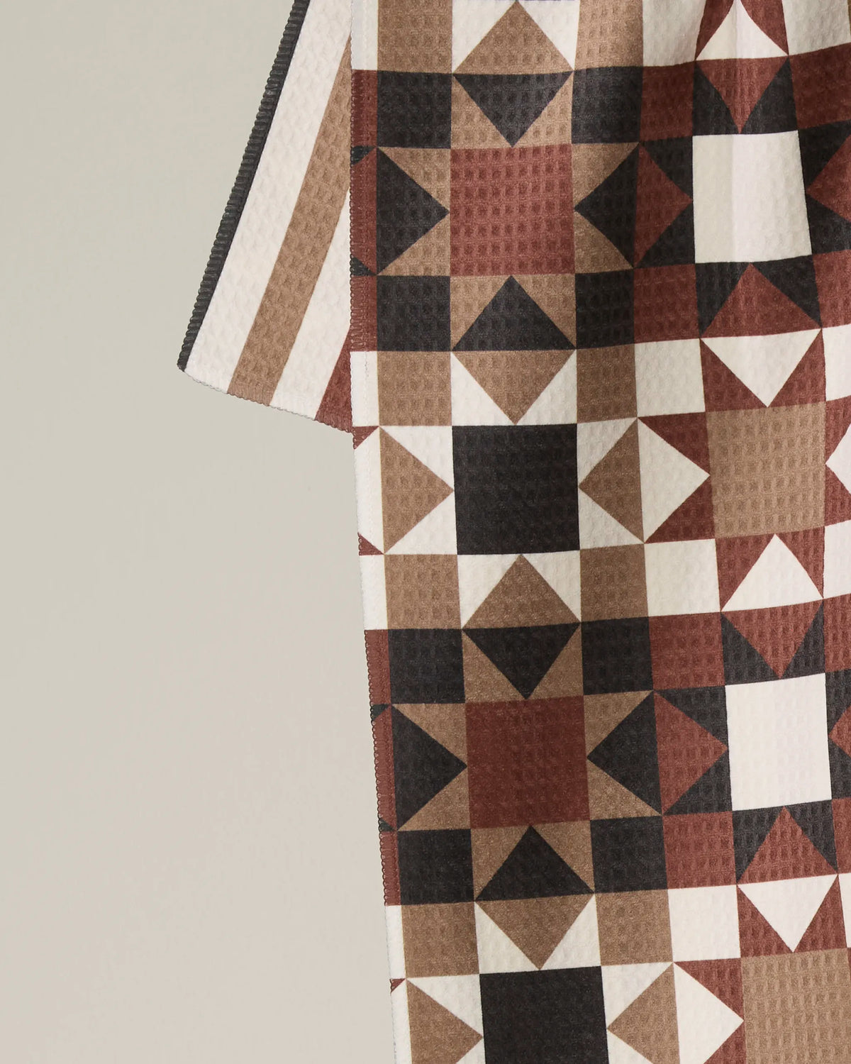 A close-up of the Geometry Quilted showcases a geometric pattern in shades of brown, black, and white, with triangles and squares arranged in a repeating grid. Crafted from materials such as a recycled kitchen towel, this unique piece by Geometry highlights creativity and sustainability. Only part of the quilt is visible against a neutral background.