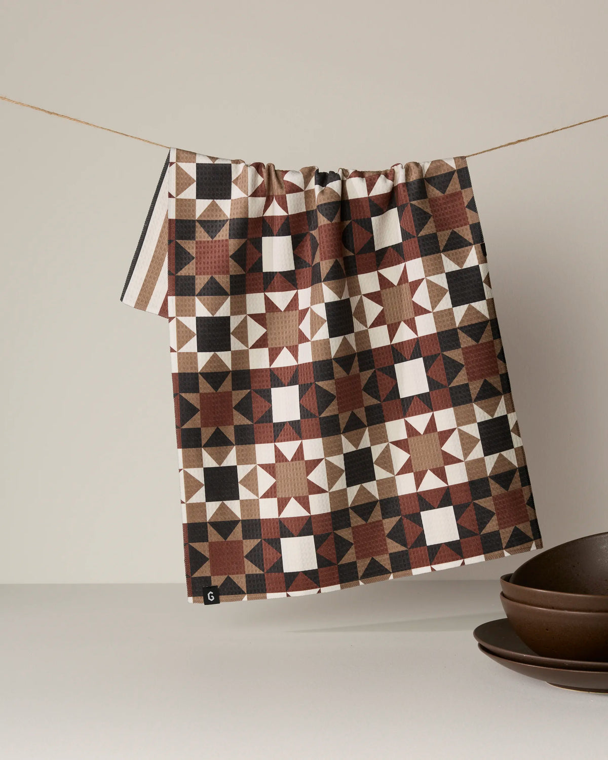 A quick-drying, double-sided Quilted tea towel by Geometry, featuring a geometric design in brown, black, and white, hangs on a string above a table set with brown ceramic bowls.
