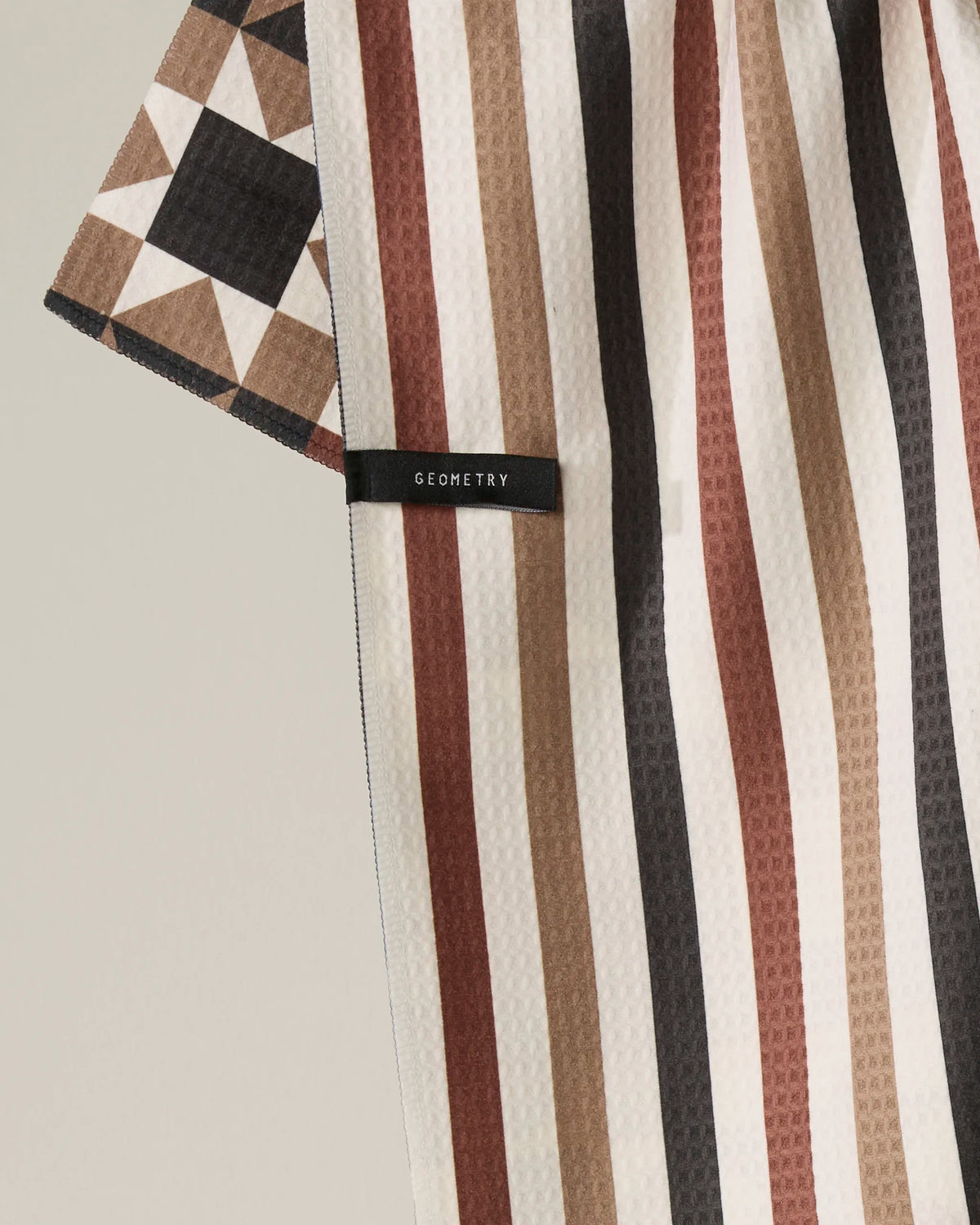 A double-sided tea towel from Geometry called “Quilted,” featuring vertical stripes in cream, brown, beige, and taupe. The corner displays a geometric triangle pattern in coordinating colors, and a black label reads “Geometry.”.