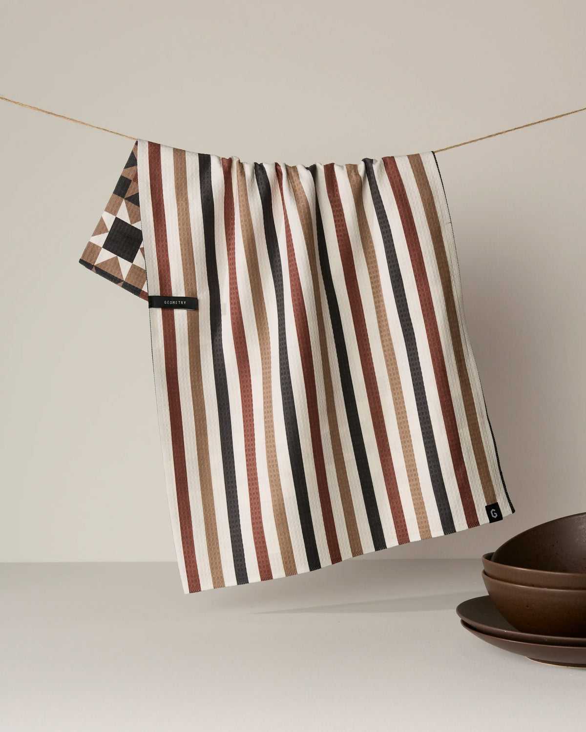 A quick-drying Geometry Quilted kitchen towel in brown, black, and cream stripes hangs on a string next to stacked brown bowls against a neutral background.