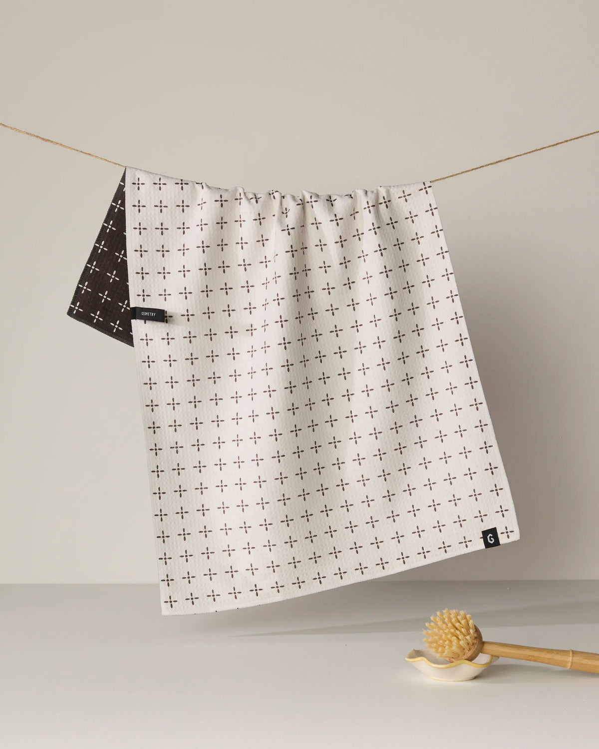 The Quinn recycled tea towel by Geometry, featuring a white double-sided design with a subtle small plus sign pattern, hangs on a string while a dark brown towel peeks out from behind. Designed for quick-drying convenience in your kitchen, the Quinn towel is displayed above a wooden honey dipper resting on the white surface below.