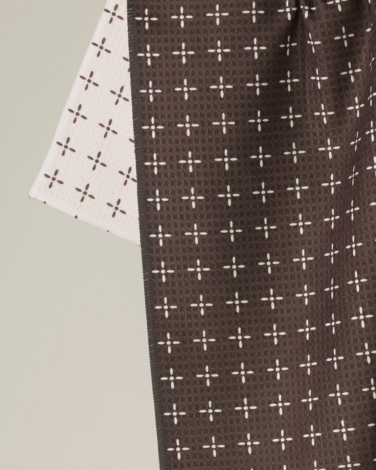 The Geometry Quinn double-sided kitchen tea towel features two overlapping pieces of fabric adorned with a repeating pattern of small white crosses and dots. The front side displays a rich brown hue, while the reverse is a pale shade—either white or light beige. Combining style and practicality, this towel is an ideal addition to any modern kitchen.