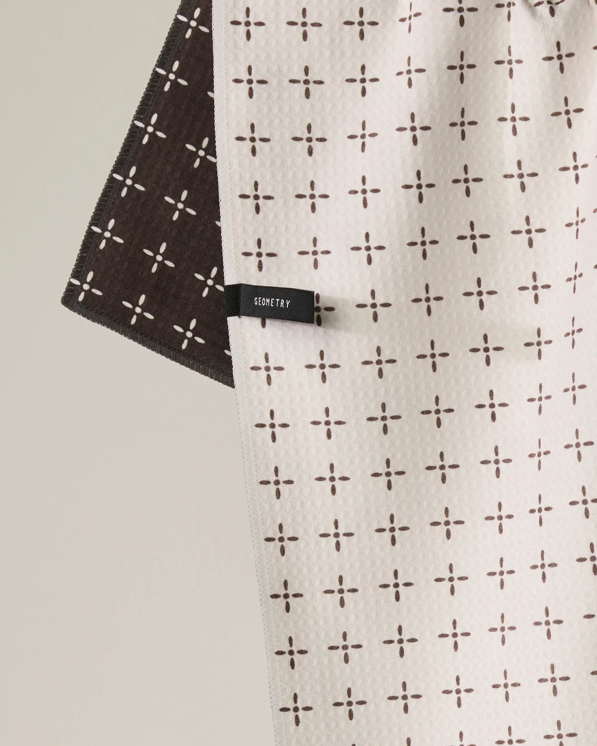 A beige and brown Double-Sided Kitchen Tea Towel called “Quinn” by Geometry hangs on display with a grid of small cross patterns visible on both sides. A black tag labeled GEOMETRY is attached to the beige side, highlighting this quick-drying tea towel’s modern design.