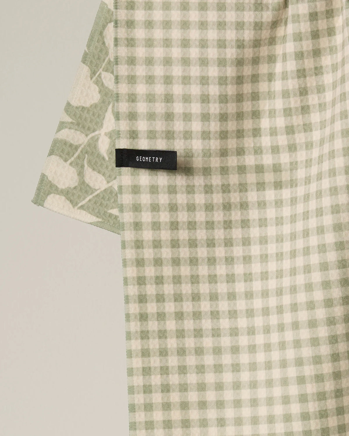 A Double-Sided Tea Towel from Geometry in the Sage Blossom design, featuring a green and white gingham pattern with a black GEOMETRY label, and a partial view of a matching leaf motif in the background.