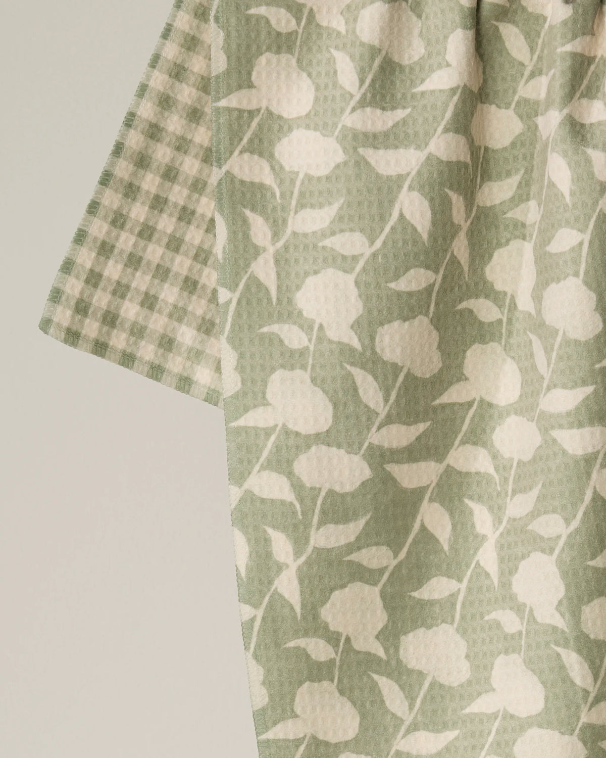 A green and white floral-patterned fabric overlays another featuring a green and white gingham check, highlighting the thoughtful double-sided design of the Geometry Sage Blossom tea towel. Crafted from recycled materials, this quick-drying tea towel is both eco-friendly and stylish—an ideal addition to any kitchen.