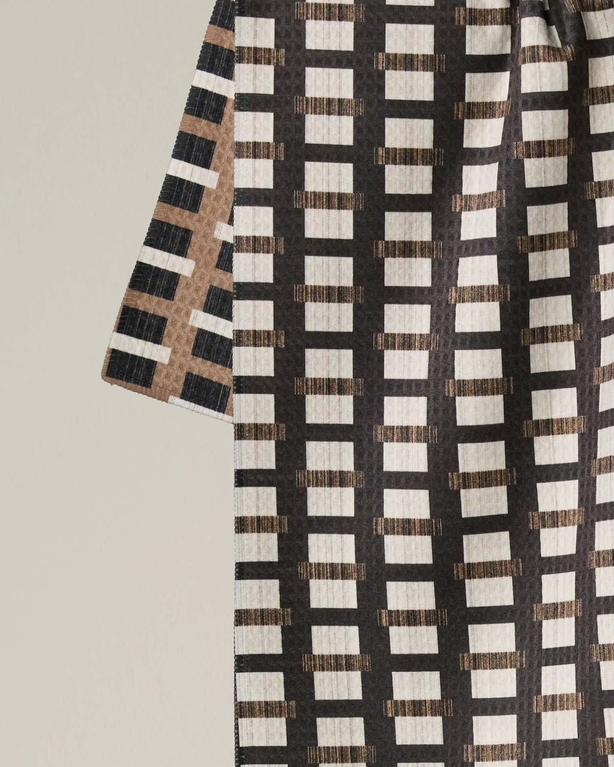 A close-up of the Geometry Terra double-sided tea towel, showcasing its geometric grid pattern with horizontal cream rectangles set against a dark background in shades of brown and black.