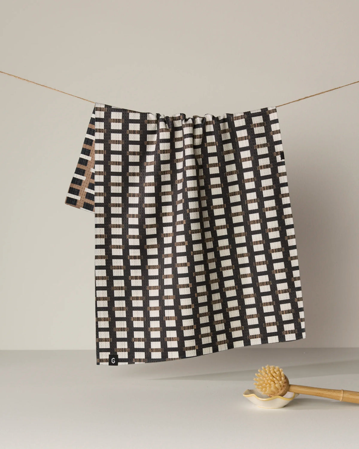 The Geometry Terra double-sided tea towel, featuring a black and white checkered design and crafted from recycled materials, hangs on a string against a plain backdrop. Below the towel, a honey dipper rests on the surface.