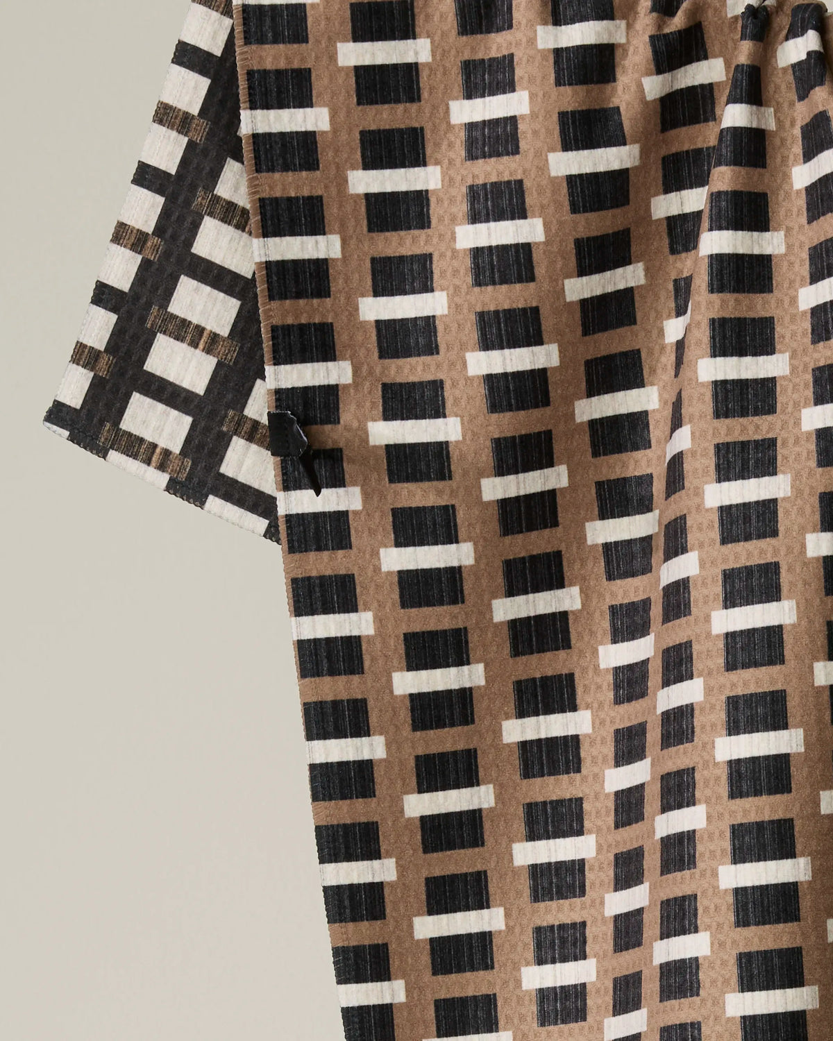 A close-up of the Geometry Terra double-sided tea towel showcases its geometric pattern, featuring rows of horizontal black and white rectangles on a brown background. The draped fabric reveals its texture and neatly stitched edge.