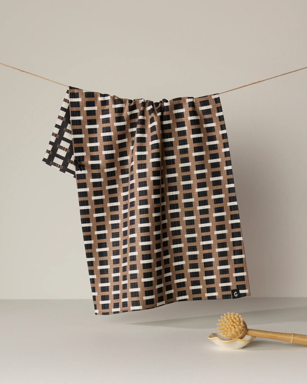 The Geometry Terra tea towel, featuring a double-sided grid design in shades of brown, black, and white, hangs on a string. Below it rests a wooden massage brush on a light surface set against a neutral background.