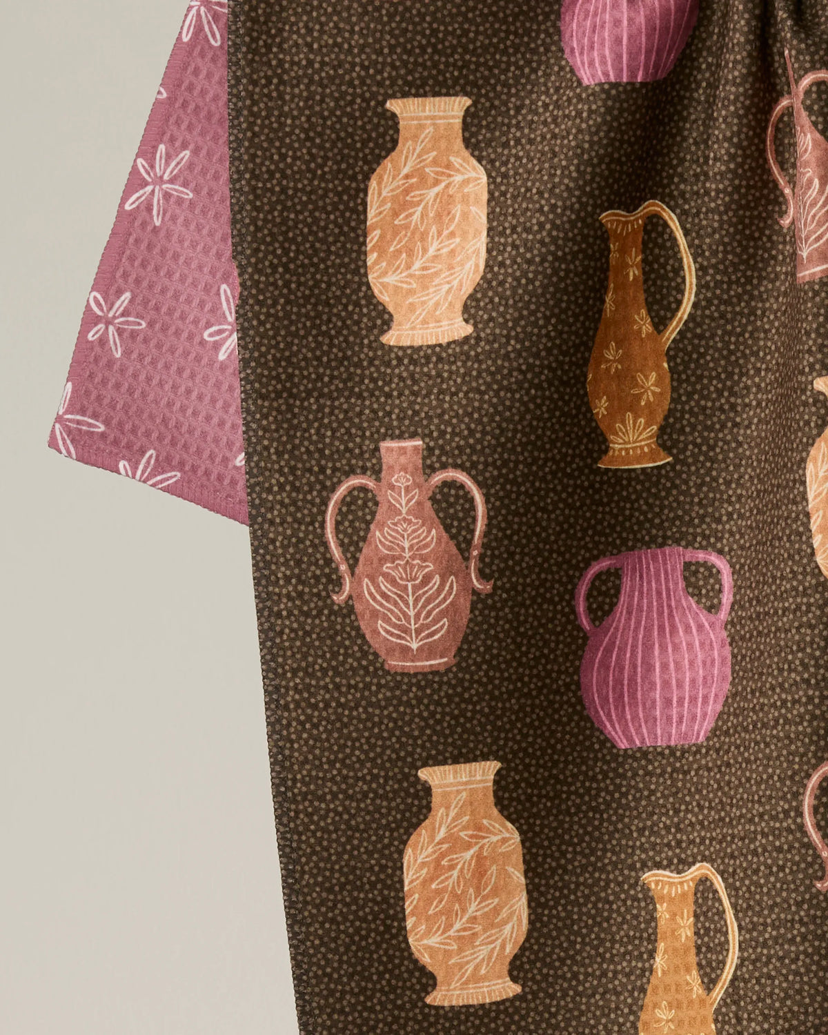 A close-up of Geometry’s “Vessels” fabric displays a pattern of illustrated vases in beige, pink, and brown tones set against a dotted dark background, with a hint of a pink patterned section—thoughtfully crafted from recycled materials for an eco-friendly finish.