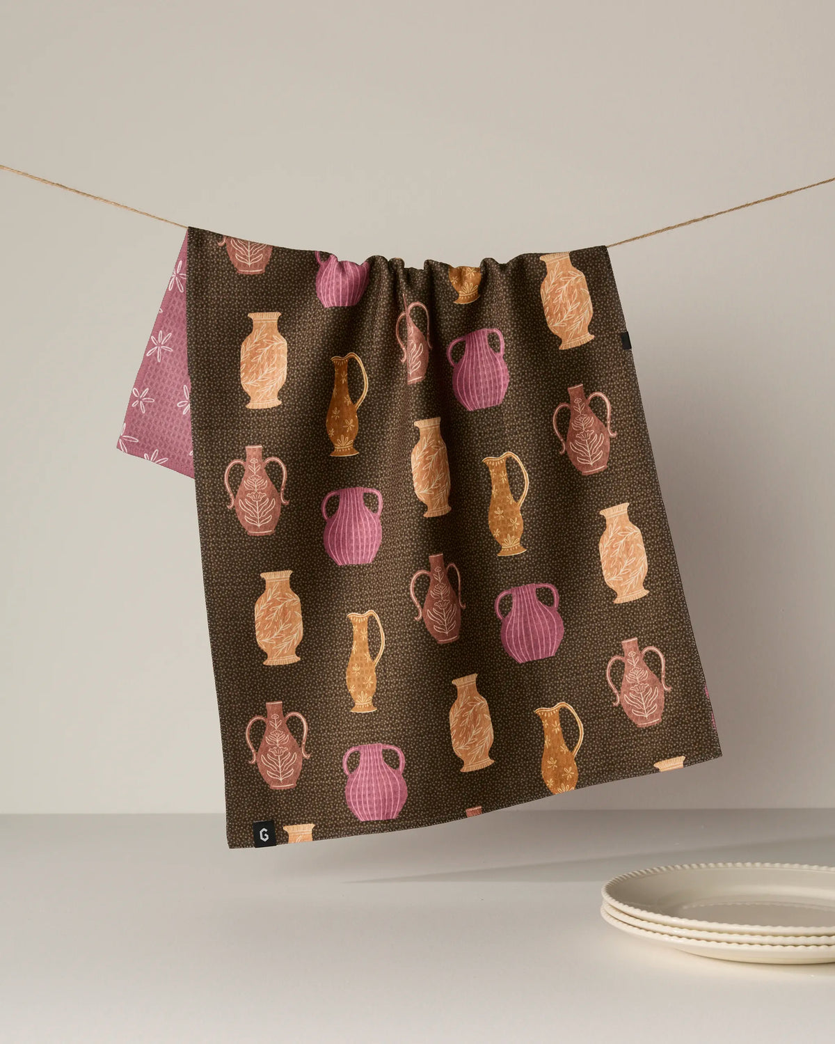 A quick-drying tea towel from Geometry, called “Vessels,” featuring brown fabric adorned with pink and orange pottery designs, hangs on a string. The edge of a white plate and a glimpse of the towel’s pink-patterned underside are visible against a simple, light background.
