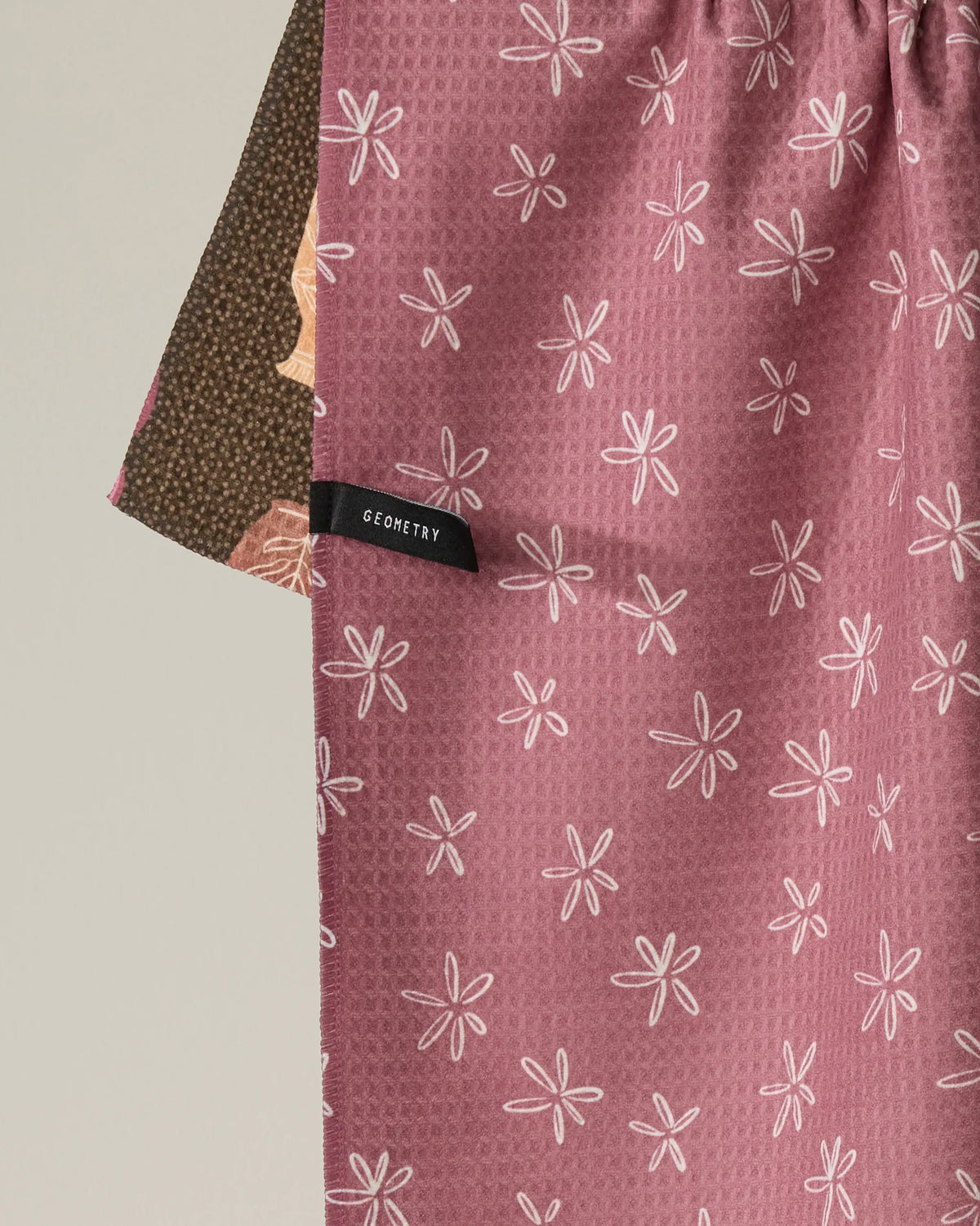A close-up of the Vessels double-sided tea towel from Geometry shows pink fabric with white floral patterns partially overlapping a brown floral design. Made from recycled materials, it features a black tag with “GEOMETRY” sewn onto the pink side.