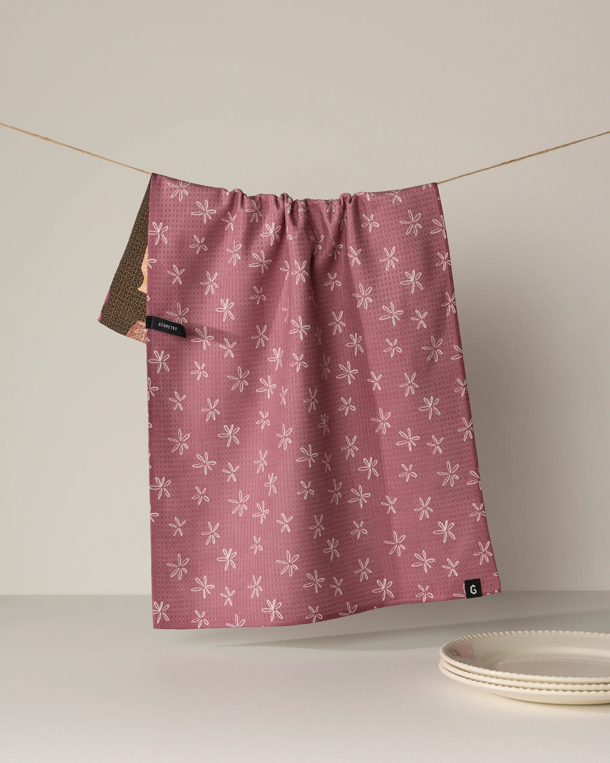 A pink Vessels cloth by Geometry, featuring a white dragonfly pattern and crafted from recycled materials, hangs on a string. Part of a brown fabric peeks from behind, and a stack of white plates sits on the table below. The background is plain and neutral.