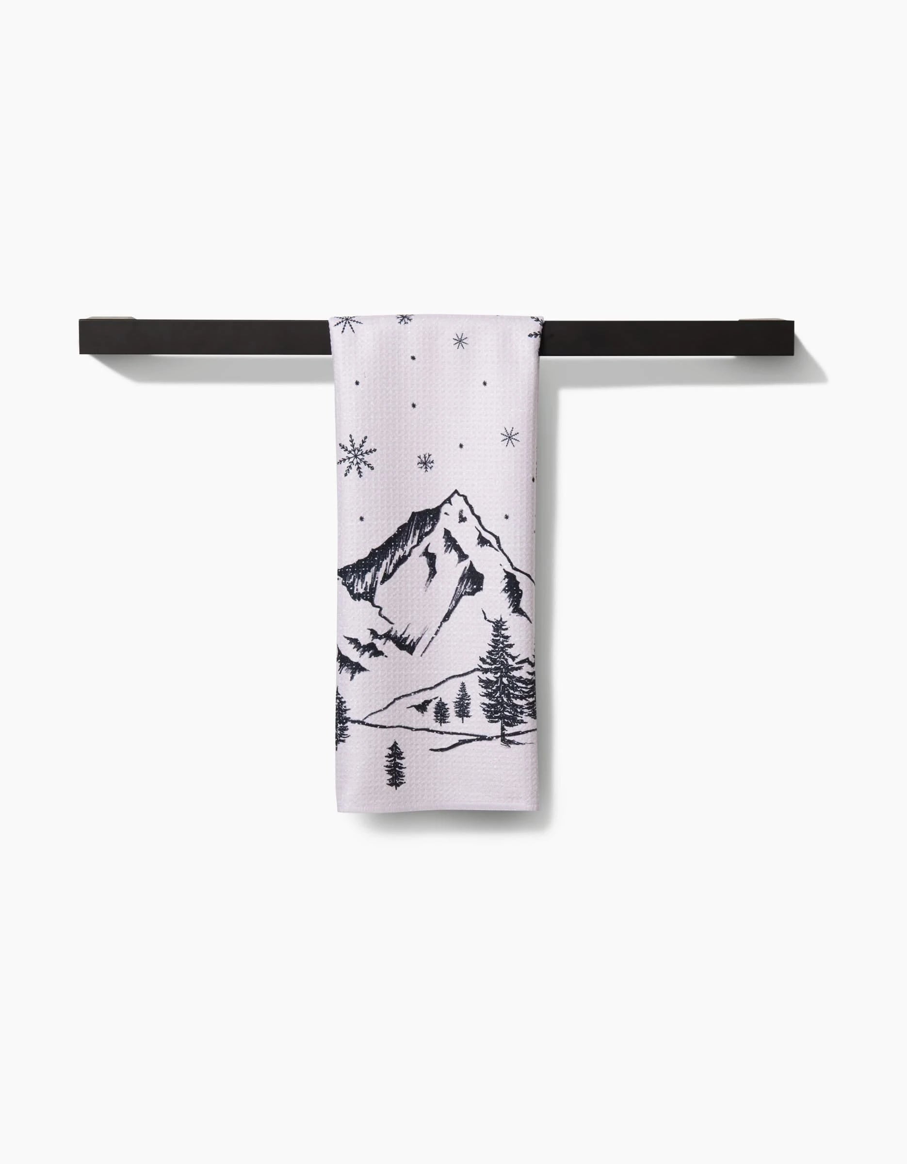 The Winter Wonderland by Becki Owens, a white ultra-absorbent towel with black mountain, pine tree, and star-like designs, is folded over a sleek black towel bar against a plain white background.