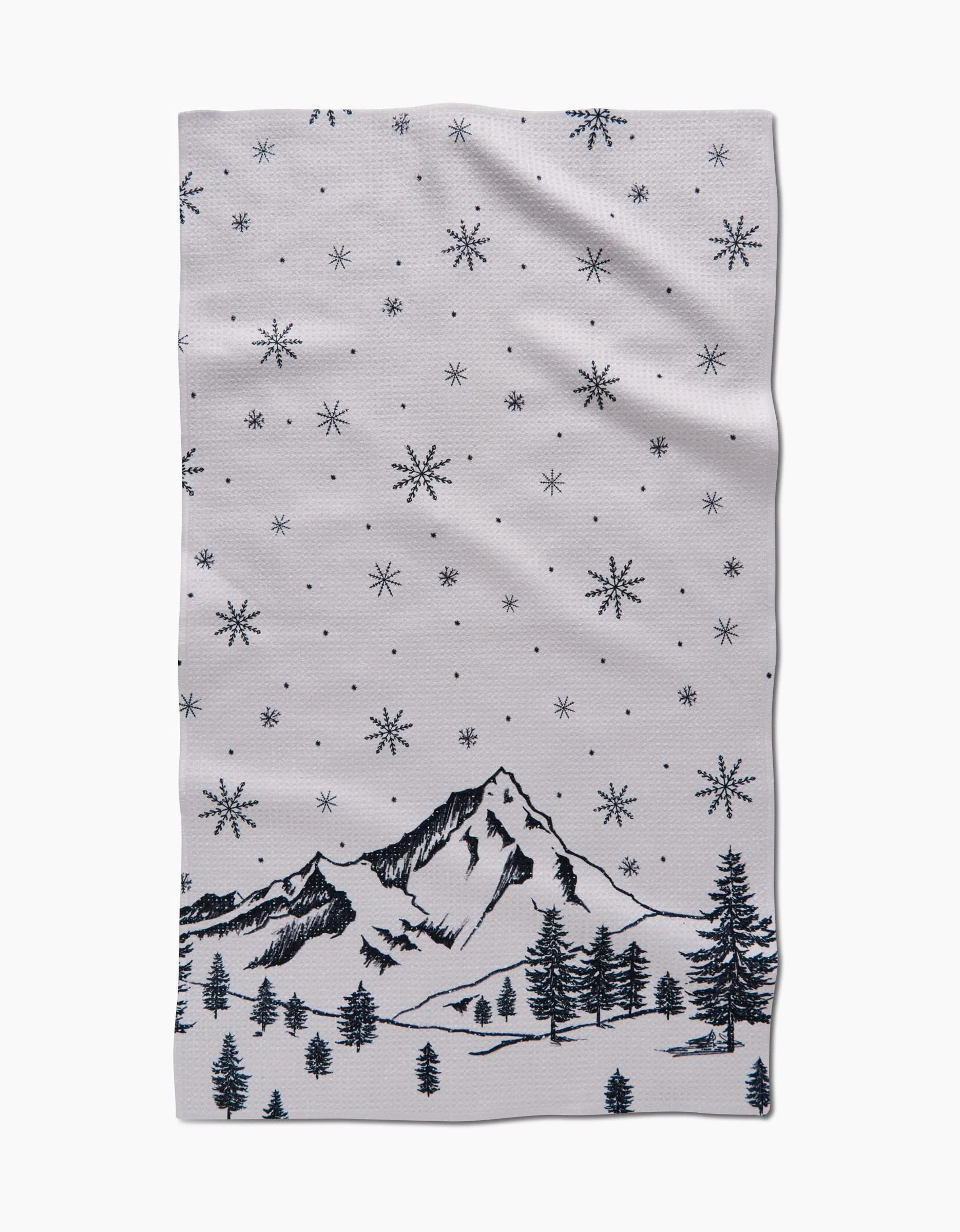 The Winter Wonderland by Becki Owens is a white recycled kitchen towel featuring black line art of snow-covered mountains, pine trees, and falling snowflakes.
