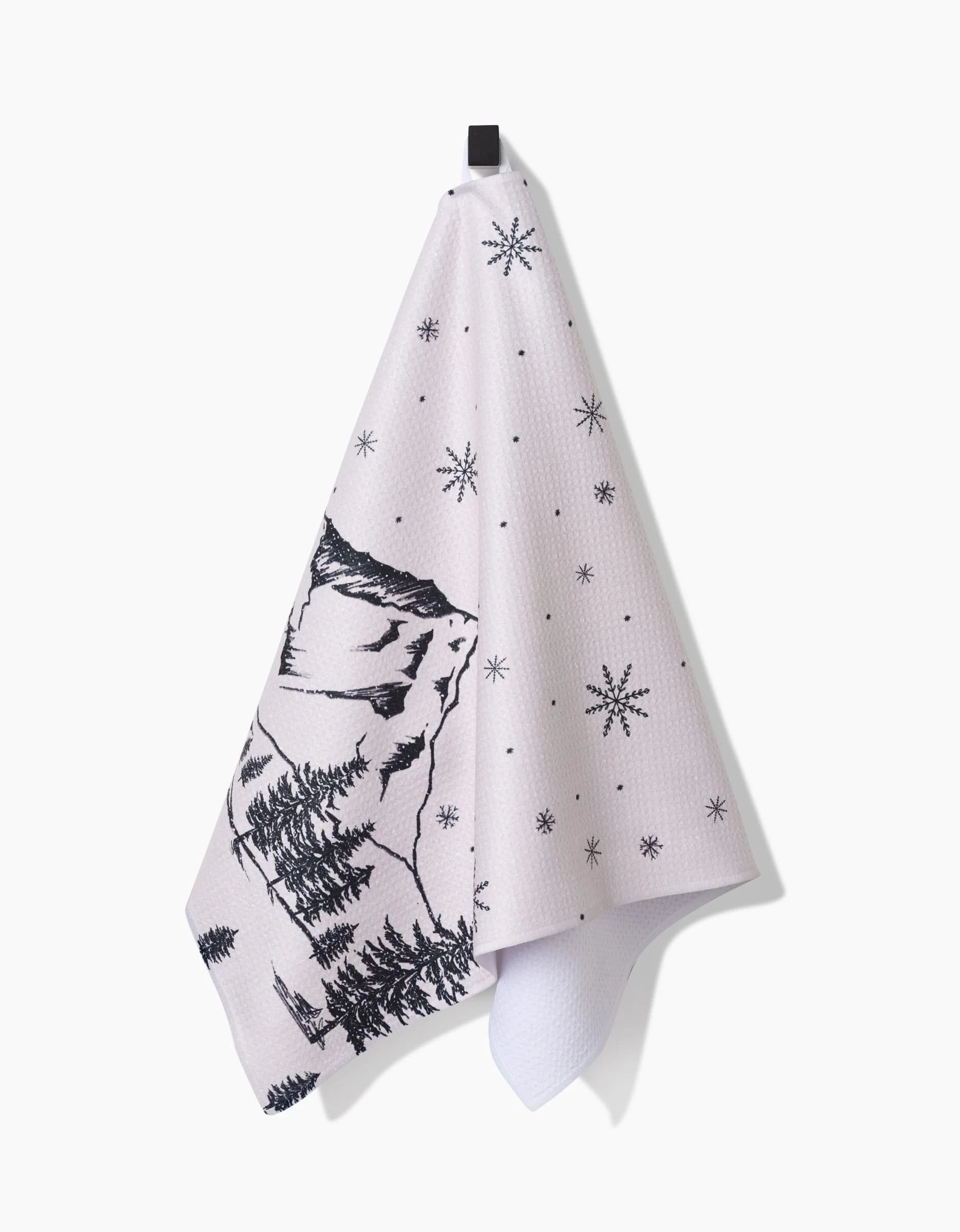 The Winter Wonderland recycled kitchen towel by Becki Owens features black line art of a forested mountain with star or snowflake patterns, shown hanging on a black hook against a plain white background.