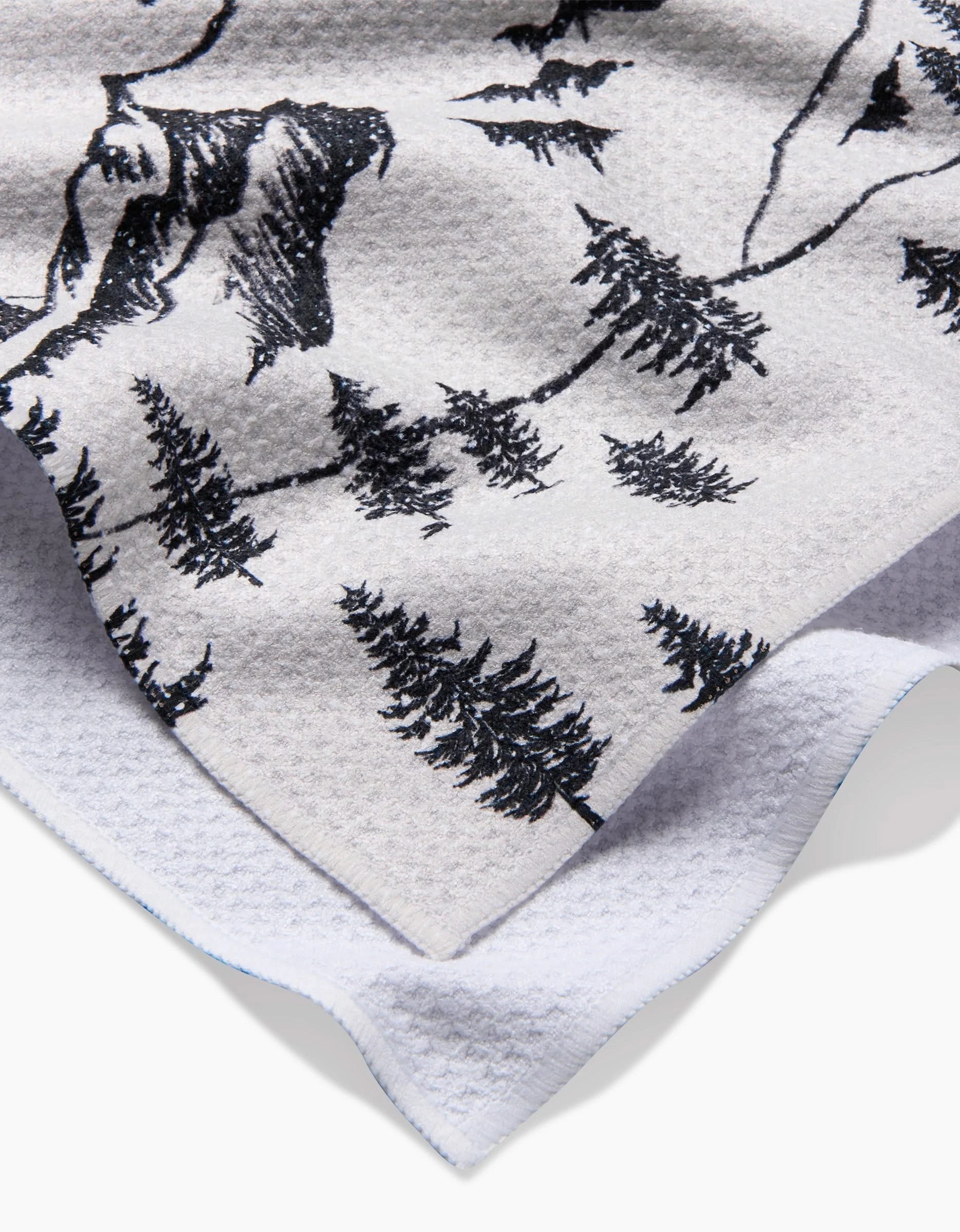 A close-up of Becki Owens’ The Winter Wonderland tea towel shows its textured white fabric and black pine tree mountain design. Ultra-absorbent, the towel is slightly wrinkled, with a folded corner revealing its reverse side.