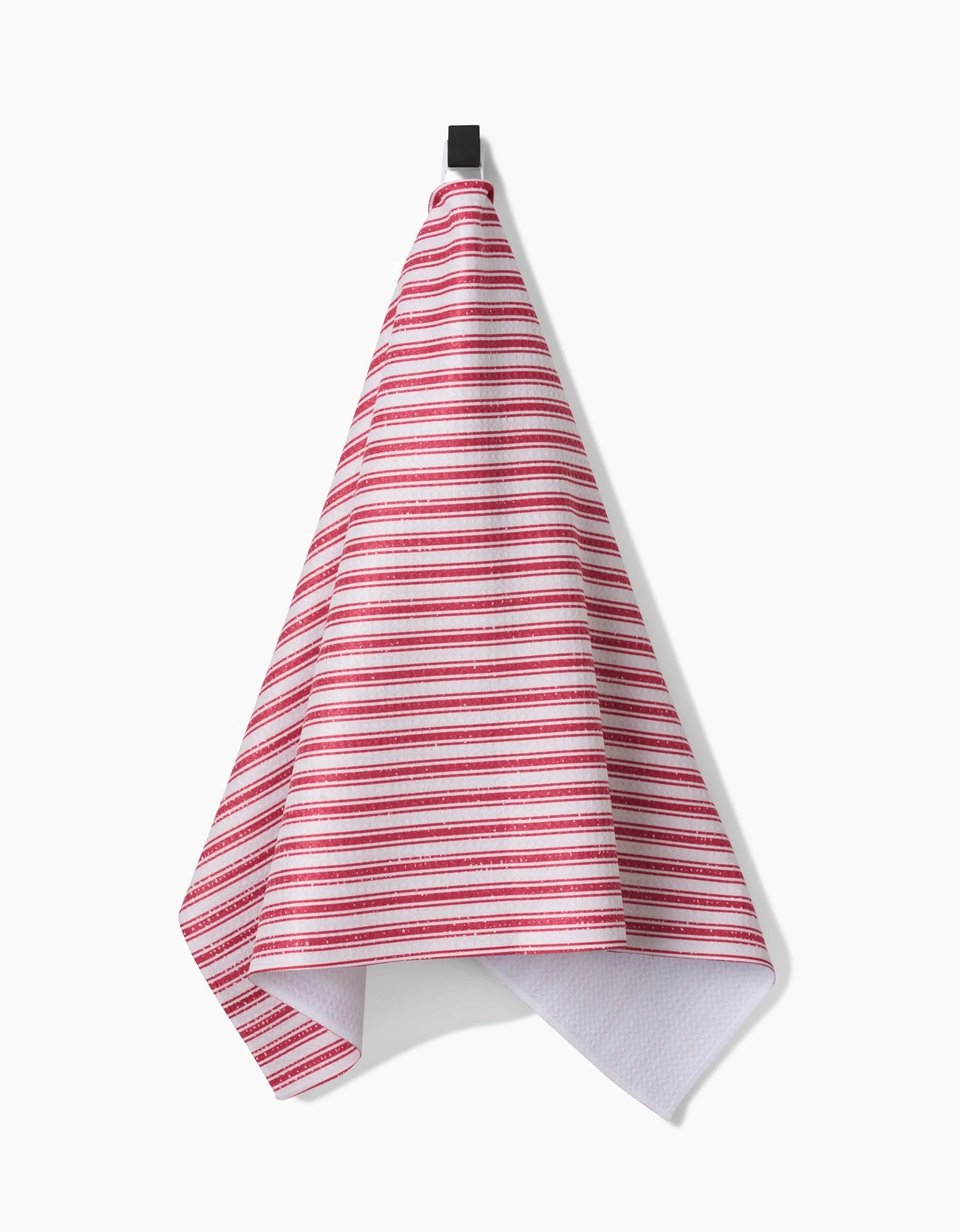 A Red Ticking tea towel by Becki Owens, made from recycled materials, hangs on a black hook against a plain white background.