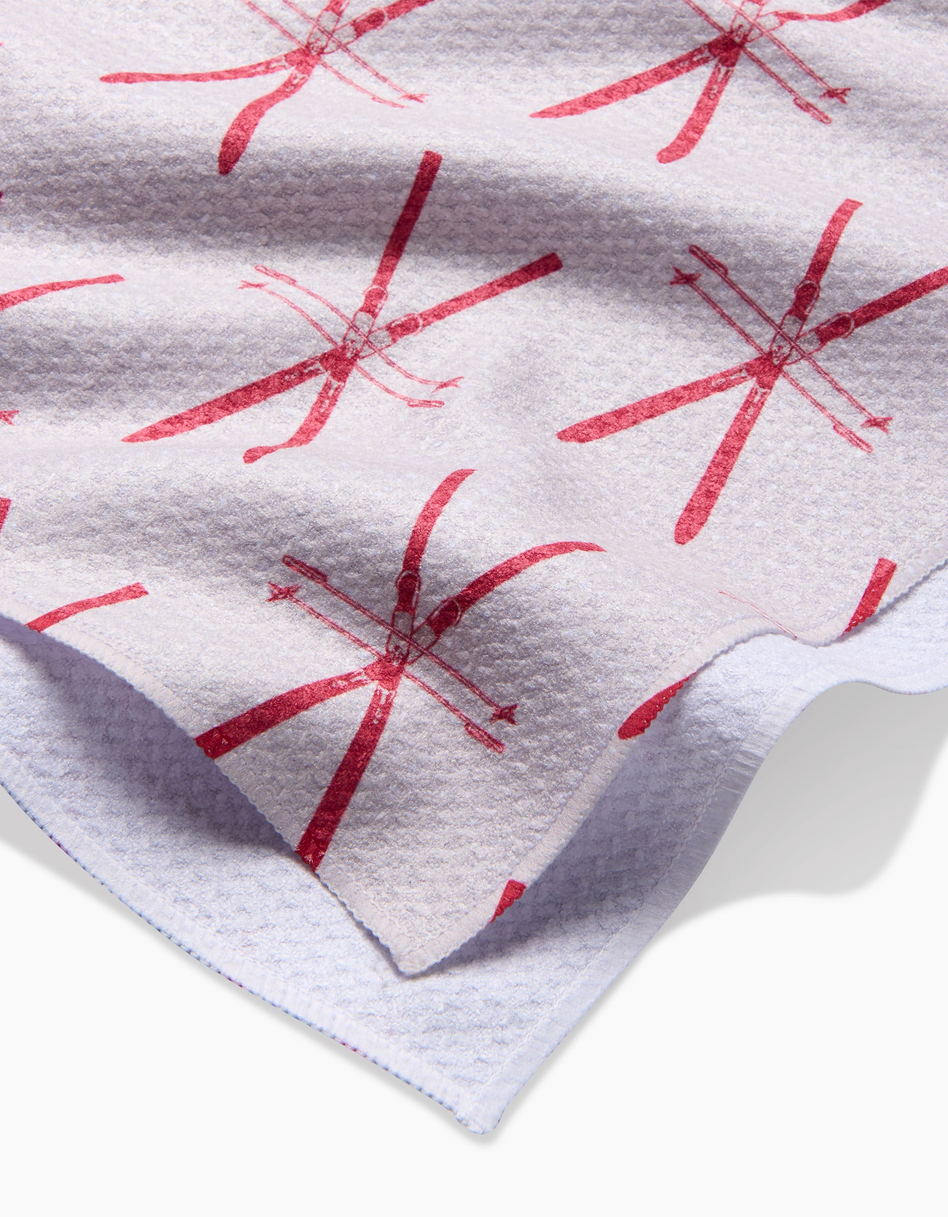 A close-up of the Vintage Ski tea towel by Becki Owens shows its white Geoweave fabric with red crossed skis and ski poles. The folded corner reveals the soft, waffle-like, eco-friendly texture.