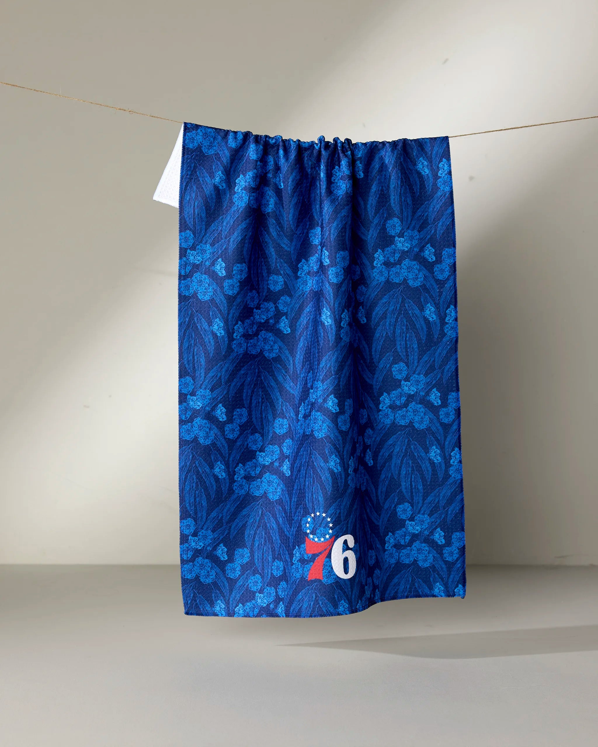 A blue sustainable kitchen towel called "76ers Floral" from NBALAB x Geometry hangs on a clothesline, showcasing a leafy pattern and the iconic red, white, and blue 76ers logo—a tribute to the Philadelphia 76ers.