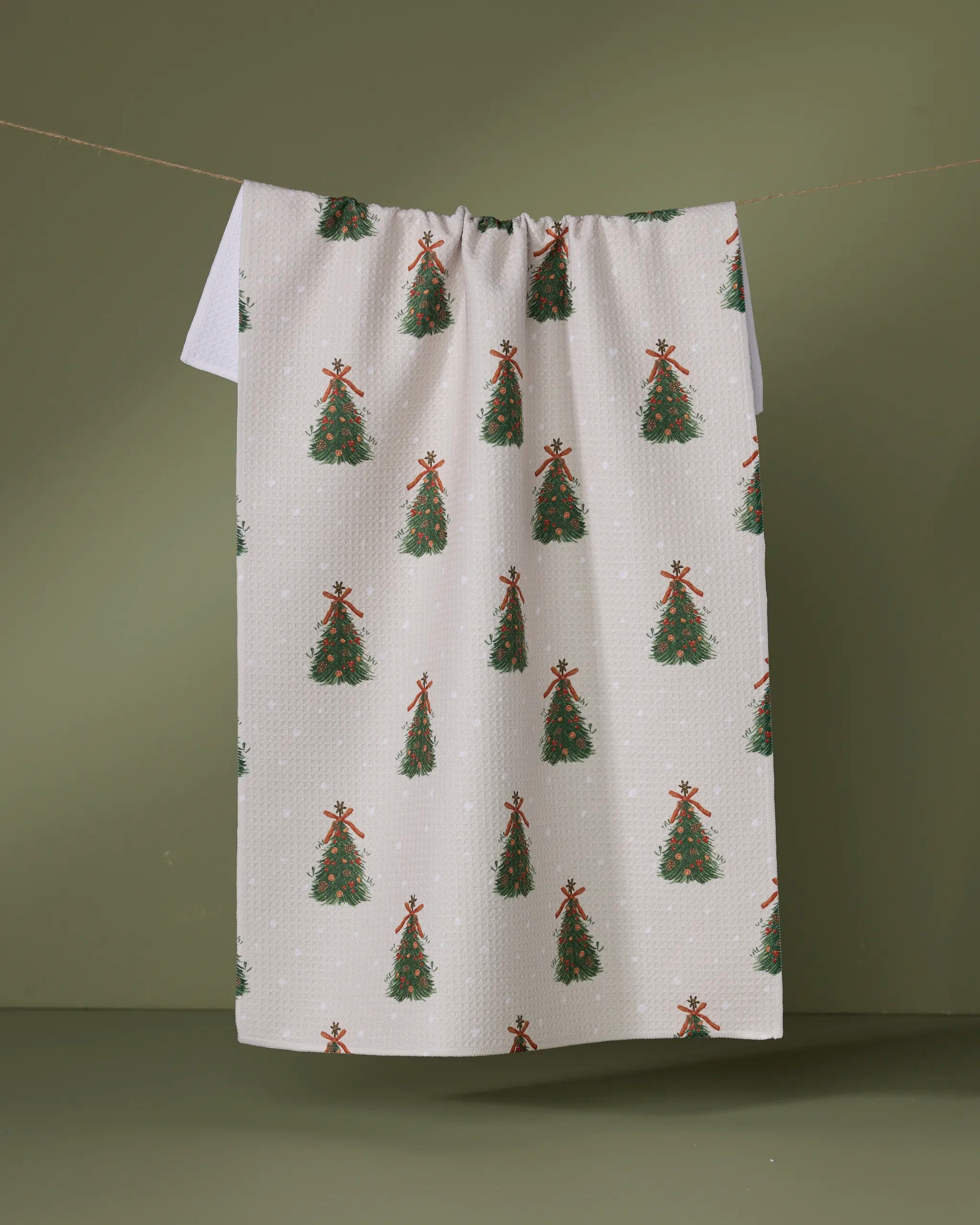 The Geometry Arbolitos white kitchen towel, made from recycled materials and featuring a repeating red-bowed Christmas tree pattern, hangs on a clothesline with an olive green background.