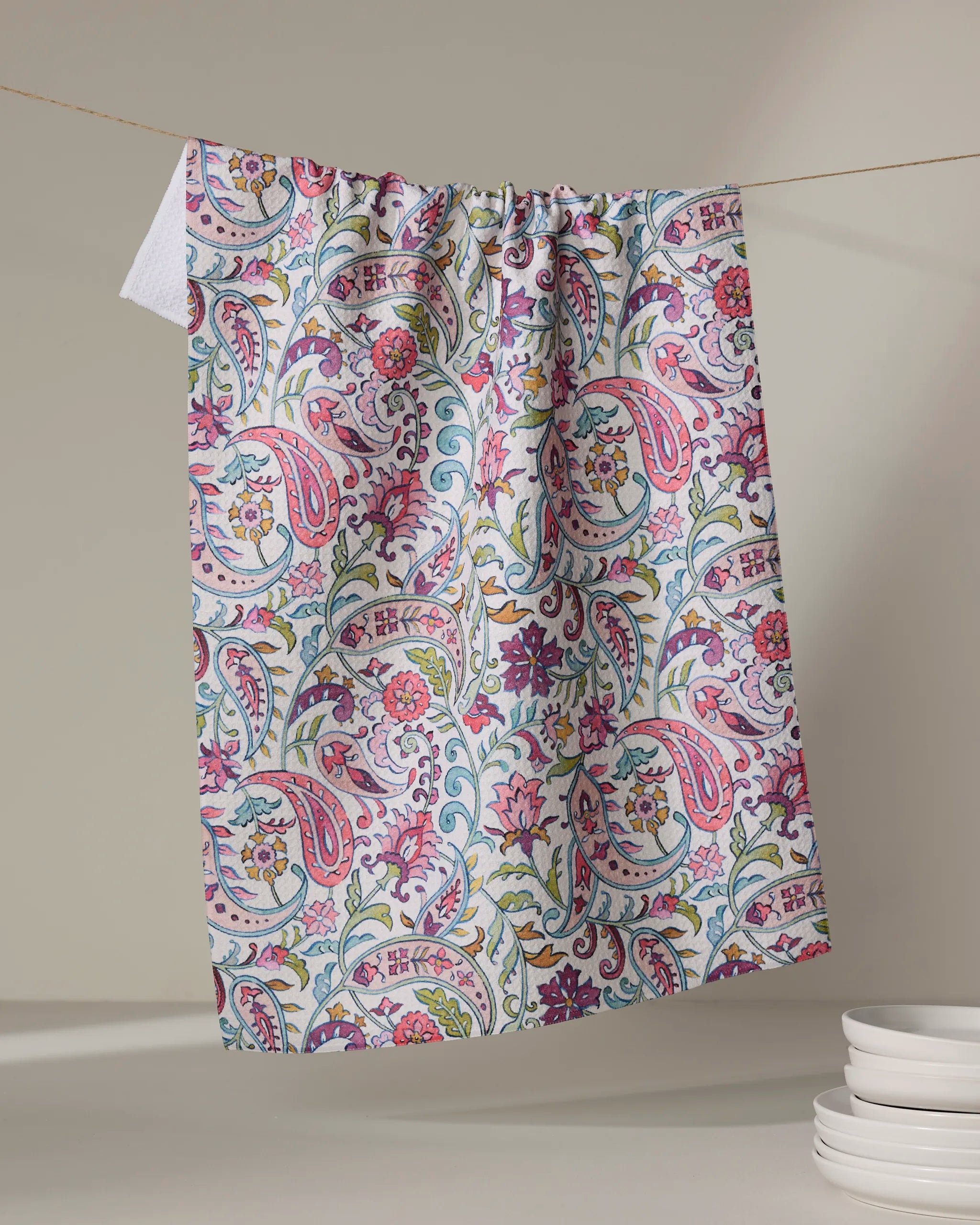 The Bazaar kitchen tea towel by Maayan Alper-Swan features a vibrant pink, green, and blue floral paisley pattern. Made from recycled materials, it brightens kitchens as it hangs near stacked white dishes against a neutral backdrop.